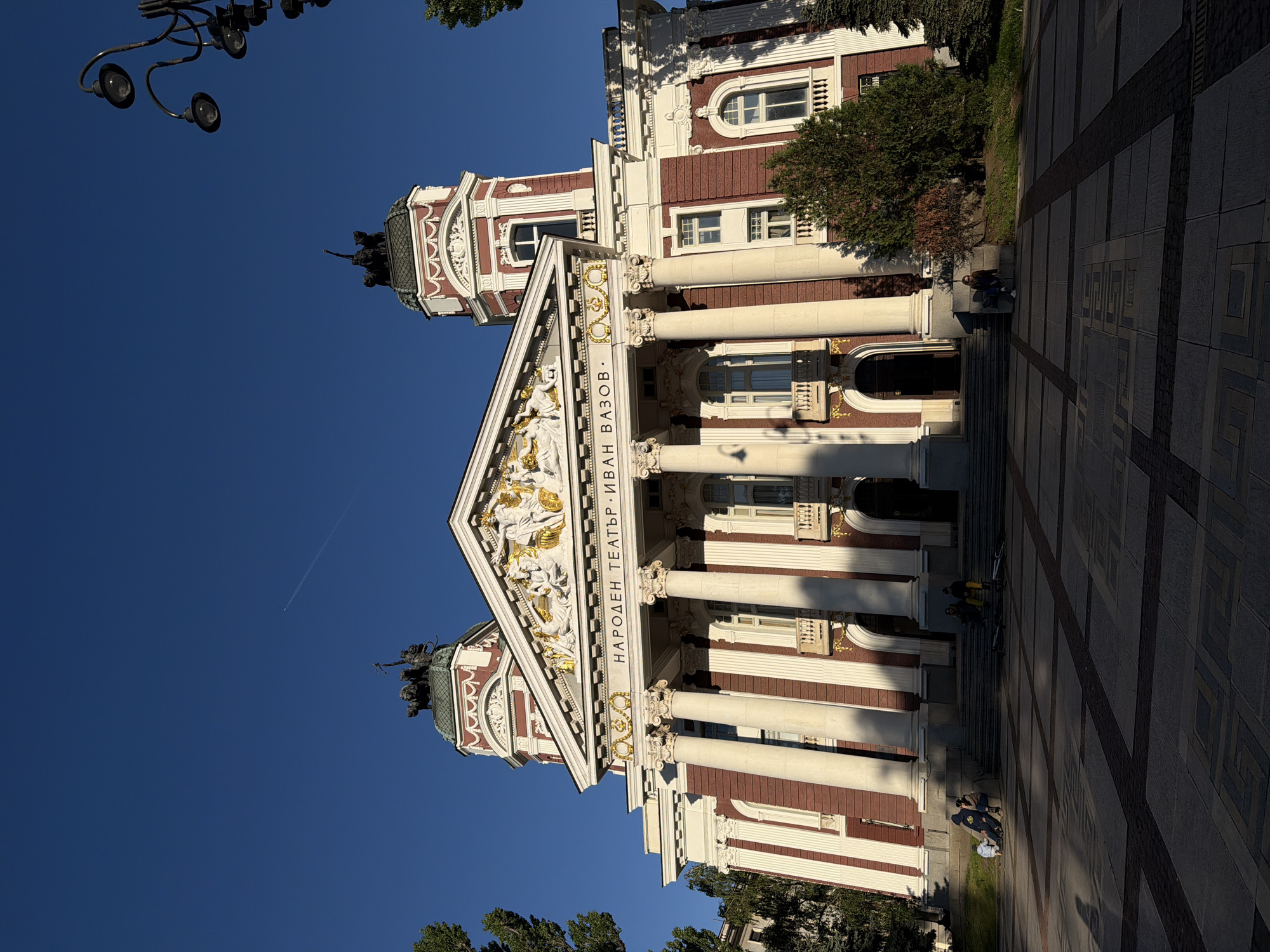 The image captures the Ivan Vazov National Theatre in Sofia, Bulgaria, showcasing a building with white columns and a red brick facade. The theater features a triangular pediment adorned with golden sculptures. A statue sits atop the building on either side of the roof. In the background is a clear blue sky and parts of a tree on the bottom left corner. In the foreground, there is a paved area, where a few people are sitting and standing. A set of ornate lamp posts can be seen to the top left of the image.