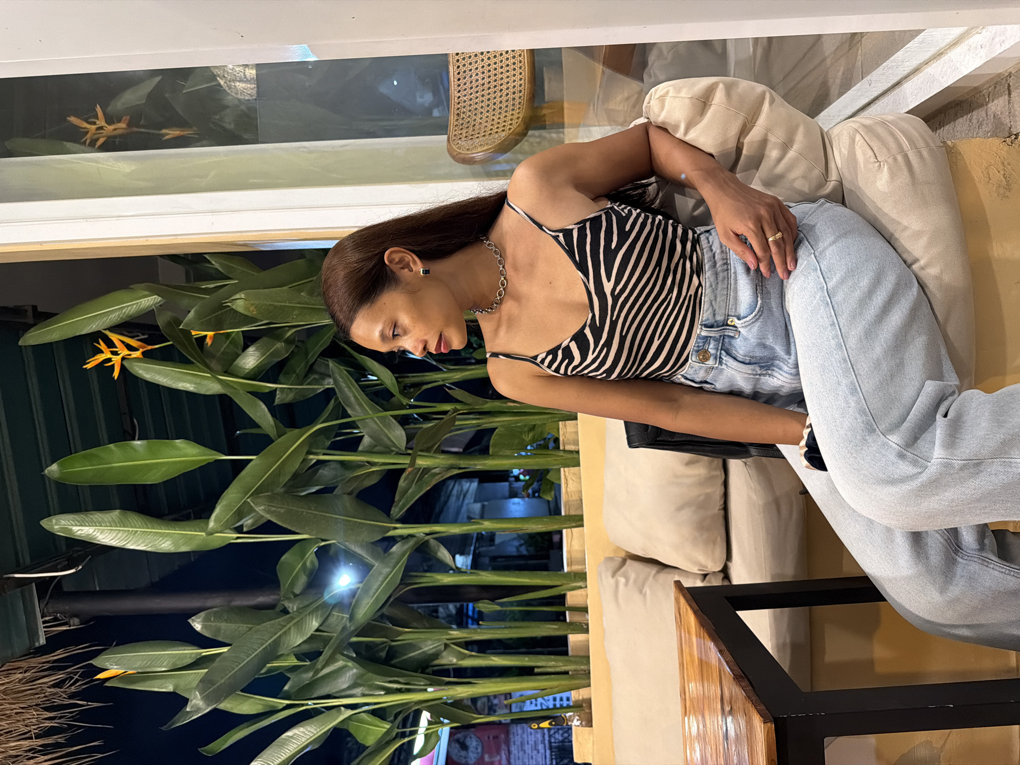 A woman sits on a light-colored cushioned couch. She is wearing a zebra print top, light blue jeans, and jewelry. The scene is located in North Kuta, Indonesia, as indicated by the vegetation and background elements. The background shows a dense cluster of green plants with long, broad leaves. An orange flower is visible among the foliage. The scene is indoors with a window or opening visible behind the plants. A portion of a wicker chair is seen in the upper part of the frame. A small table with a wooden and black frame is next to the couch. The lighting is a combination of natural and artificial. The woman appears relaxed.