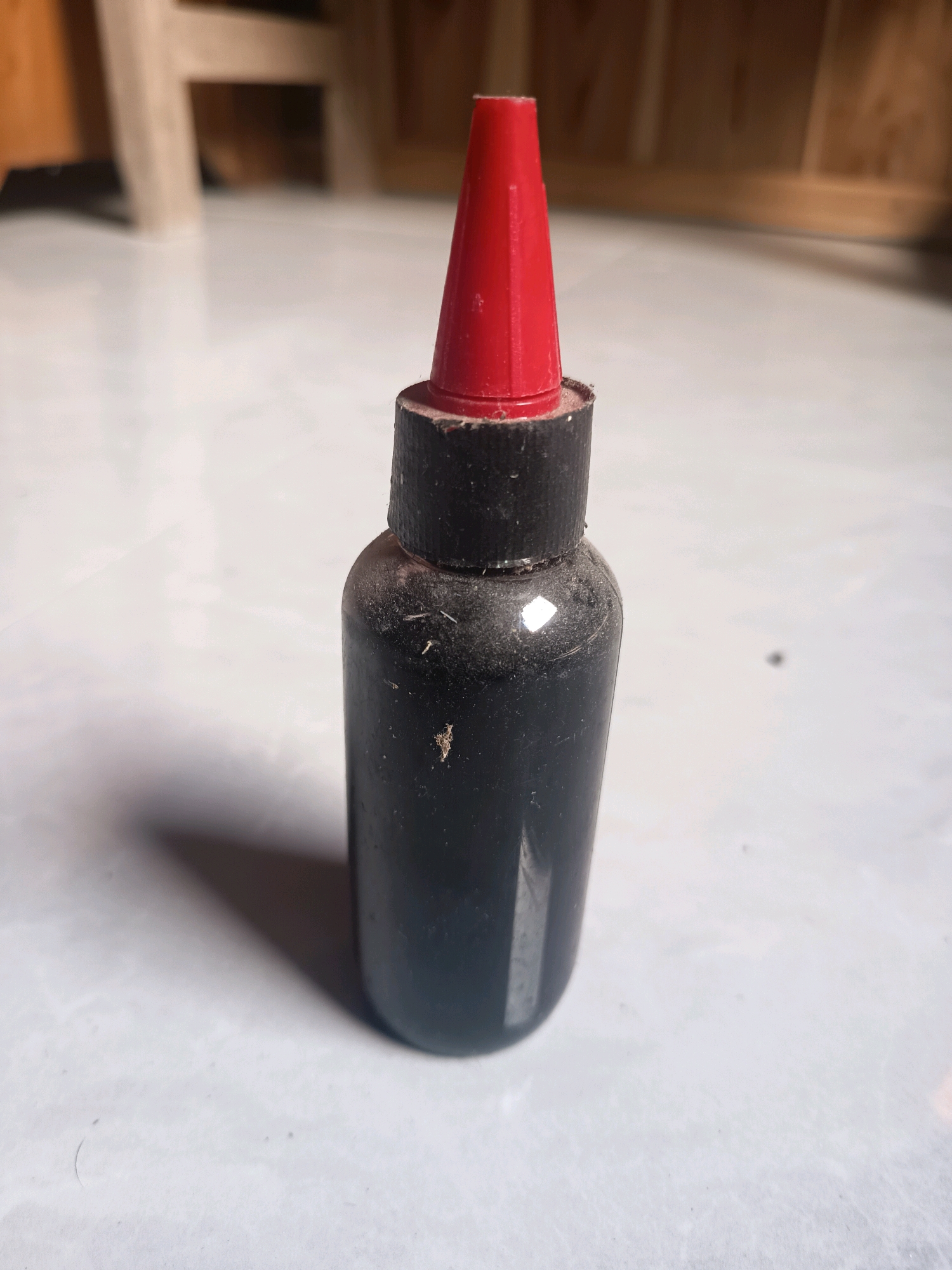 A black plastic bottle with a red tip is standing on a light-colored tiled floor in what appears to be a room in Sidorejo, Indonesia. The bottle is upright and centered in the frame, with its red cap forming a conical shape at the top. The bottle itself is dark and slightly dusty. The background shows blurred wooden furniture and a light-colored floor, suggesting an indoor setting. The lighting is somewhat dim, casting a shadow from the bottle onto the floor. There are no people or other discernible objects in the immediate vicinity of the bottle.