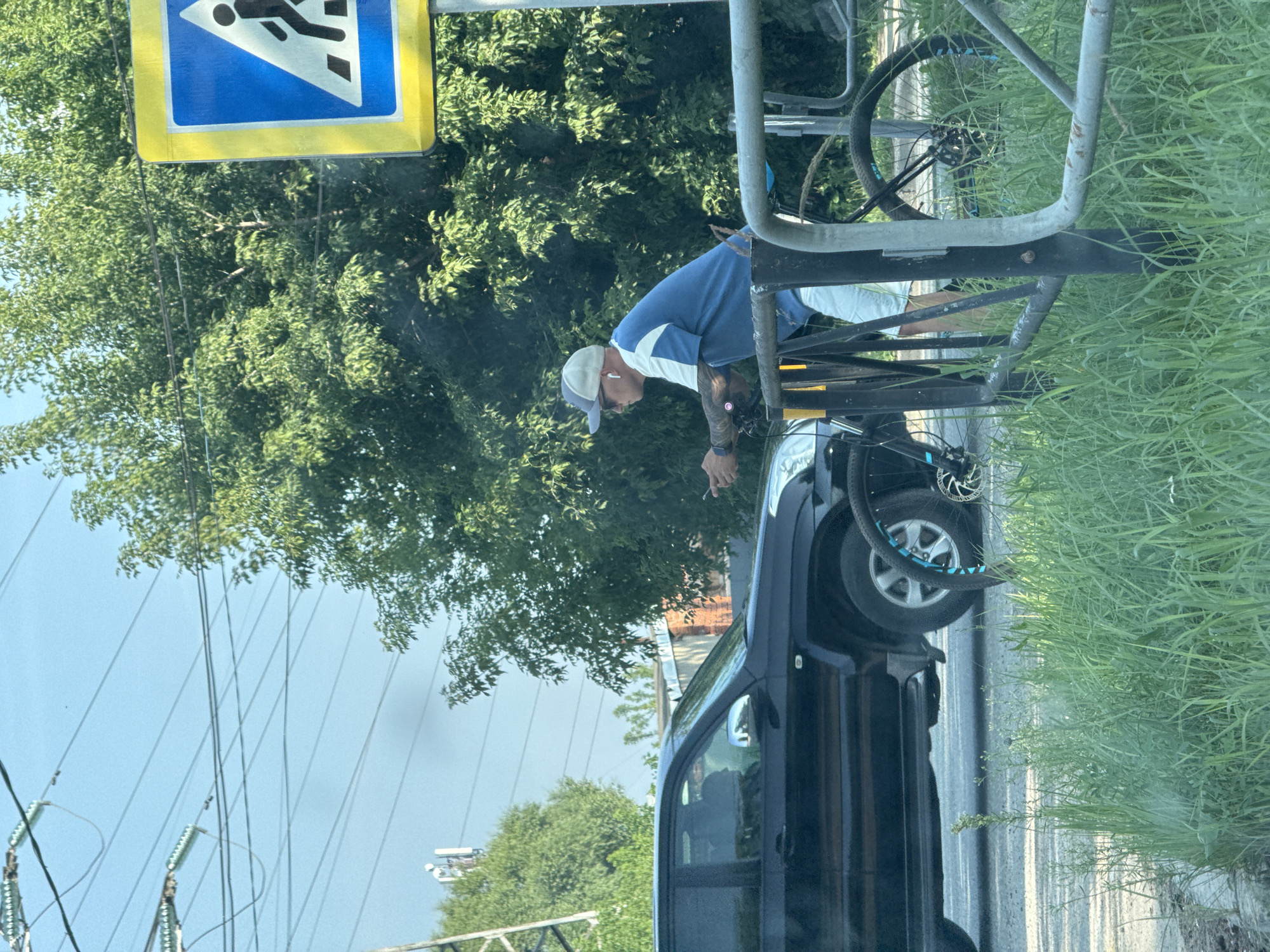 A man wearing a blue and white shirt, a baseball cap, and earbuds is leaning on a bicycle rack. He appears to be smoking. The man is standing next to a dark-colored car. In the background, there are trees and power lines. A blue and yellow pedestrian crossing sign is visible in the upper left corner of the image. The image is taken in Chaikovsky, Russia, and appears to be during the daytime, as indicated by the lighting. The overall scene suggests an outdoor environment, possibly a roadside or park.