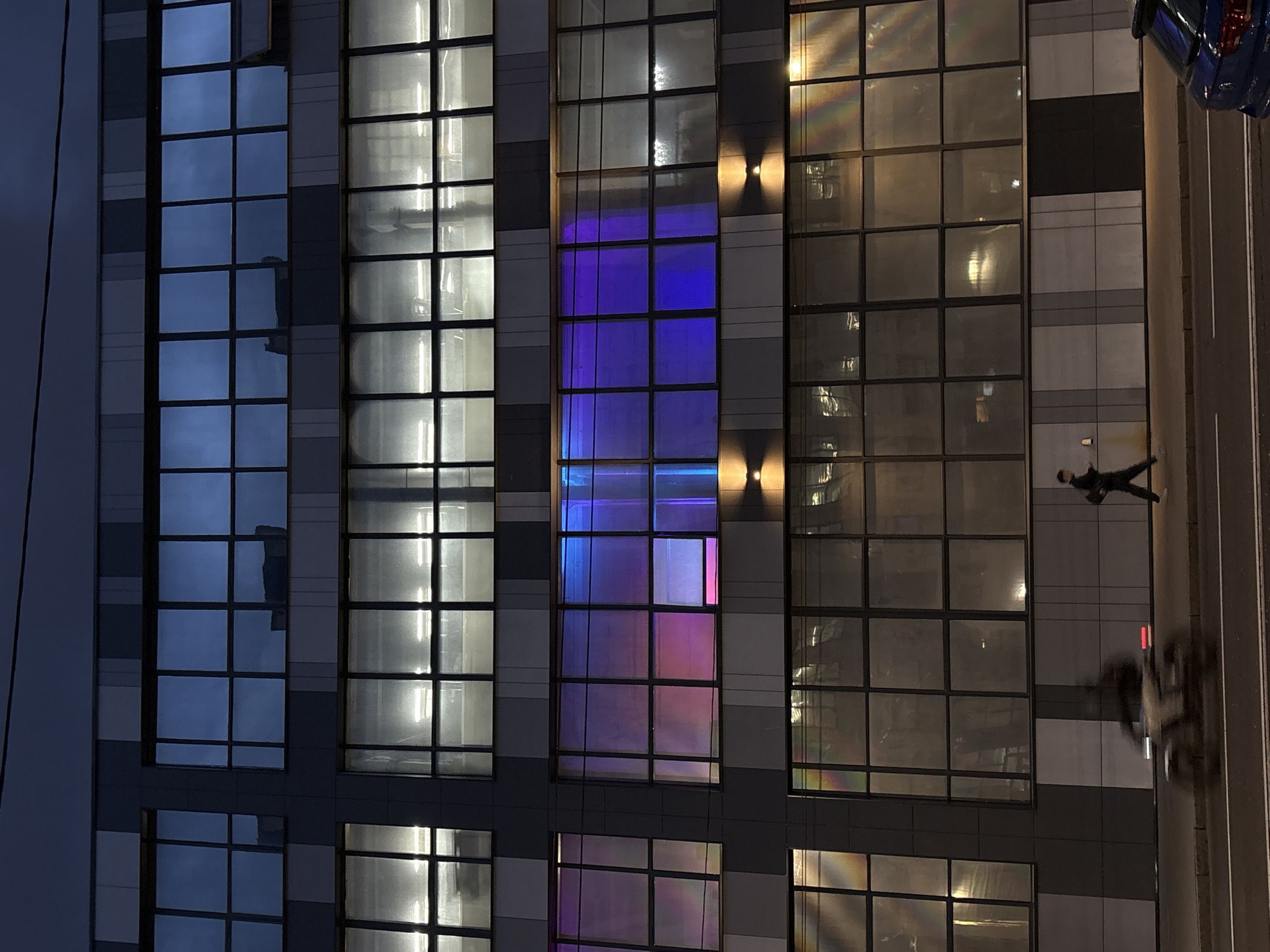 The image shows an upward view of a multi-story building with a modern glass facade, likely taken at dusk or night. The building is located near a street with cars and a pedestrian.
Here's a breakdown:
* **Building:** The building has multiple columns of windows, each with a grid-like pattern. The windows reflect various light sources, including sky light and artificial light. One column of windows reflects a purple and blue light.
* **Street:** Beside the building, there is a street with at least one car visible. A pedestrian can be seen walking on the sidewalk next to the building.
* **Environment:** The sky is dark, suggesting it is late in the day.
It is not possible to determine the exact location of the building in Minsk, Belarus, based on the image alone.