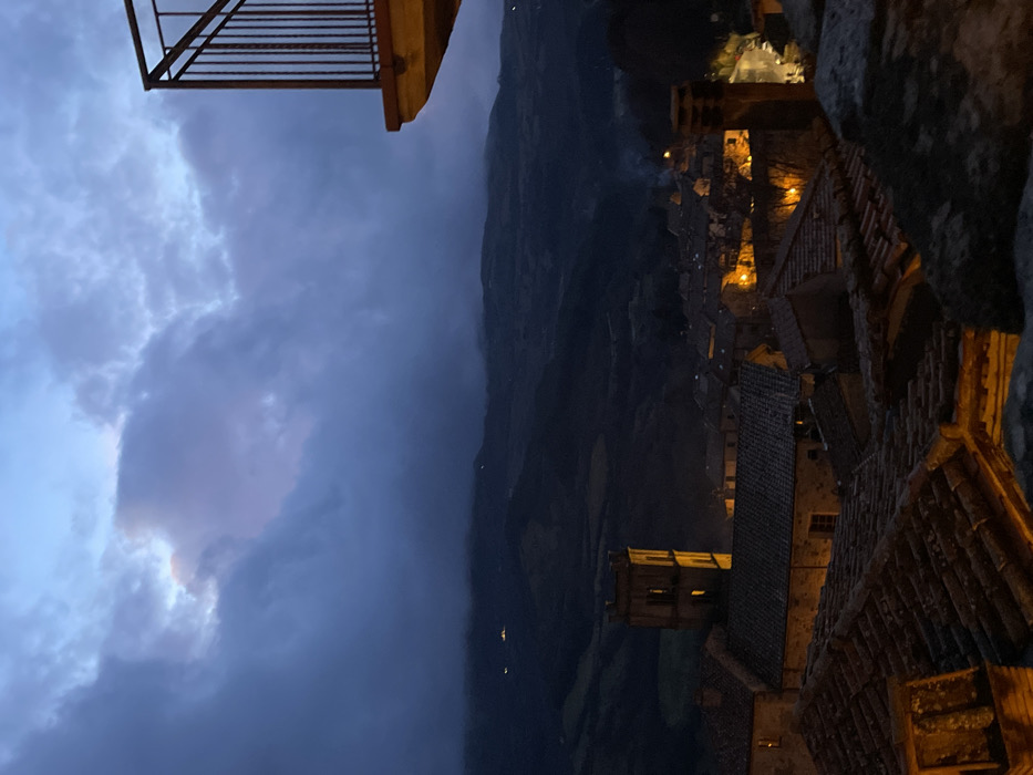 This image, best viewed rotated 90 degrees clockwise to a landscape orientation, presents a dramatic dusk or night scene over the historic town of Santa Fiora, Italy. The vantage point appears to be from a building, with a dark metal balcony railing visible in the upper foreground (top right if rotated).
The main subject is a compact cluster of old European-style buildings, characteristic of an Italian village, nestled within a valley or mountainous terrain. These stone buildings feature traditional tiled roofs and are brightly lit by numerous warm, yellow lights emanating from windows and street fixtures, casting a golden glow across the scene. A prominent square tower, possibly a church bell tower, stands out among the houses.
Beyond the illuminated town, a vast, dark mountainous landscape stretches into the background, with only a scattering of faint lights visible in the distance across the dark slopes. The sky dominates the left side of the frame (top left if rotated) and is heavily overcast with dark blue-grey clouds. Lighter, almost ethereal patches of clouds, possibly catching the last vestiges of daylight or moonlight, are visible towards the far left (top left corner if rotated).
No people are visible, and no specific activity is taking place beyond the town being lit up for the evening. The scene conveys a sense of historic charm and tranquil beauty, emphasizing the atmospheric contrast between the warmly lit village and the dark, cloudy sky and mountains. No visible text is present in the image.