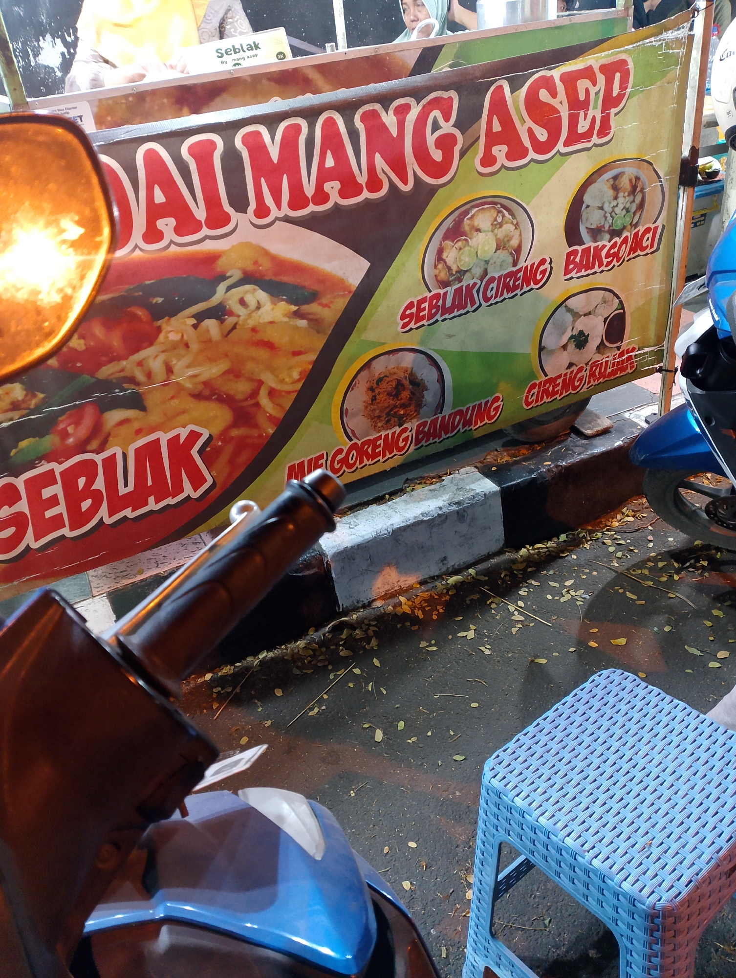 The image captures a street-level view in Magelang, Indonesia, likely at night given the artificial lighting. The focal point is a banner advertising "DAI MANG ASEP," a food vendor. The banner displays images of various Indonesian dishes including "SEBLAK CIRENG," "BAKSOACI," and "MIE GORENG BANDUNG." In the foreground, parts of a blue motorcycle are visible, including the handlebars and front fairing. A blue plastic stool sits on the ground near the motorcycle. The ground is paved and covered with scattered fallen leaves. The background shows the vendor's stall with a person partially visible behind it, wearing a headscarf.