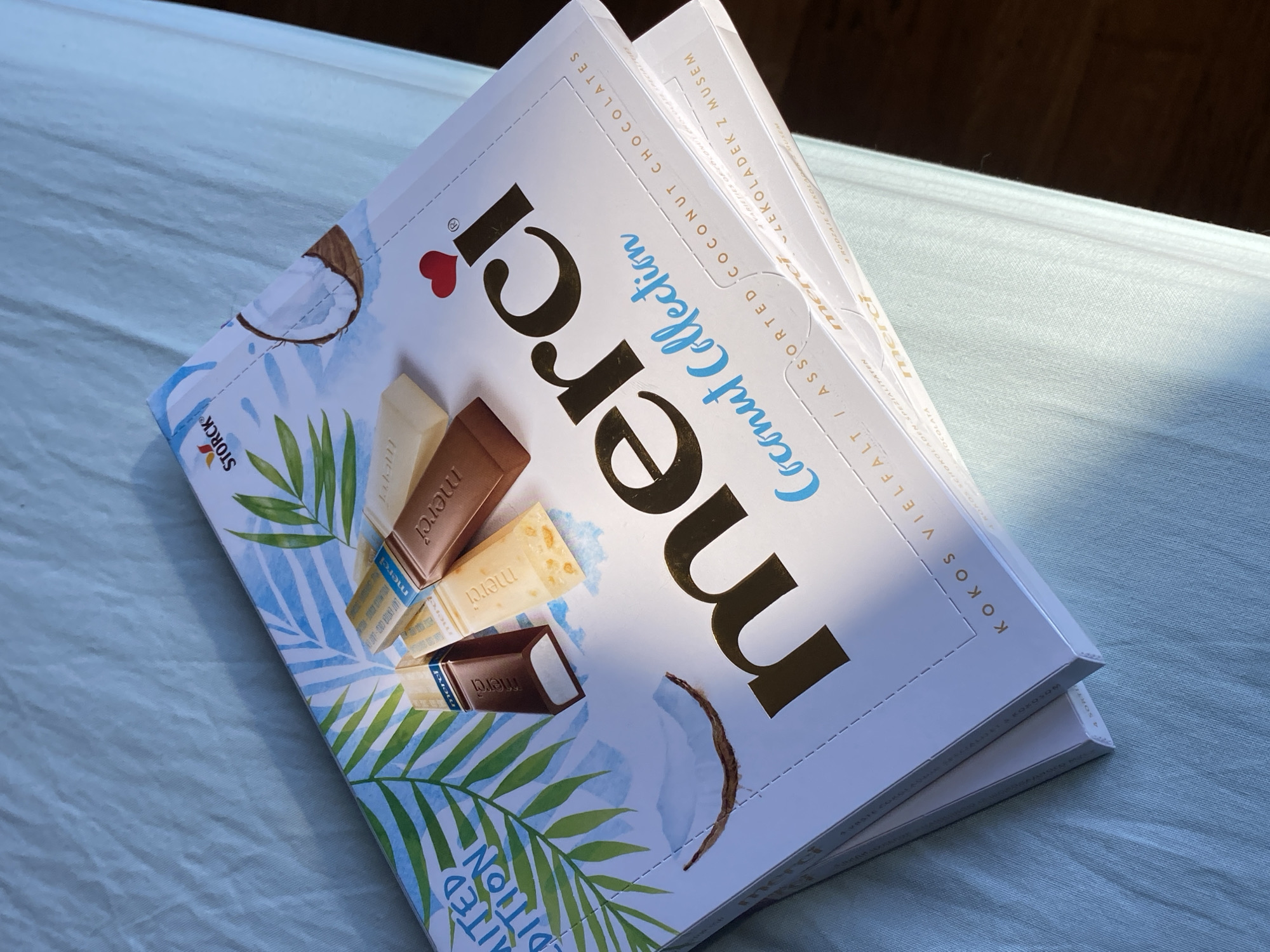 A pile of Merci Coconut Collection chocolate boxes is displayed on a light blue, textured surface, possibly a tablecloth or bedspread, under soft lighting. The top box features tropical leaf illustrations, a coconut half, and the Merci logo in gold lettering. Small individual chocolate bars are visible on top of the main box. The lower box is partially obscured but also appears to be a Merci chocolate box. The overall scene suggests a product display or an arrangement of chocolates. The provided context states the location is Wrocław, Poland, but this is not visually evident in the image itself.