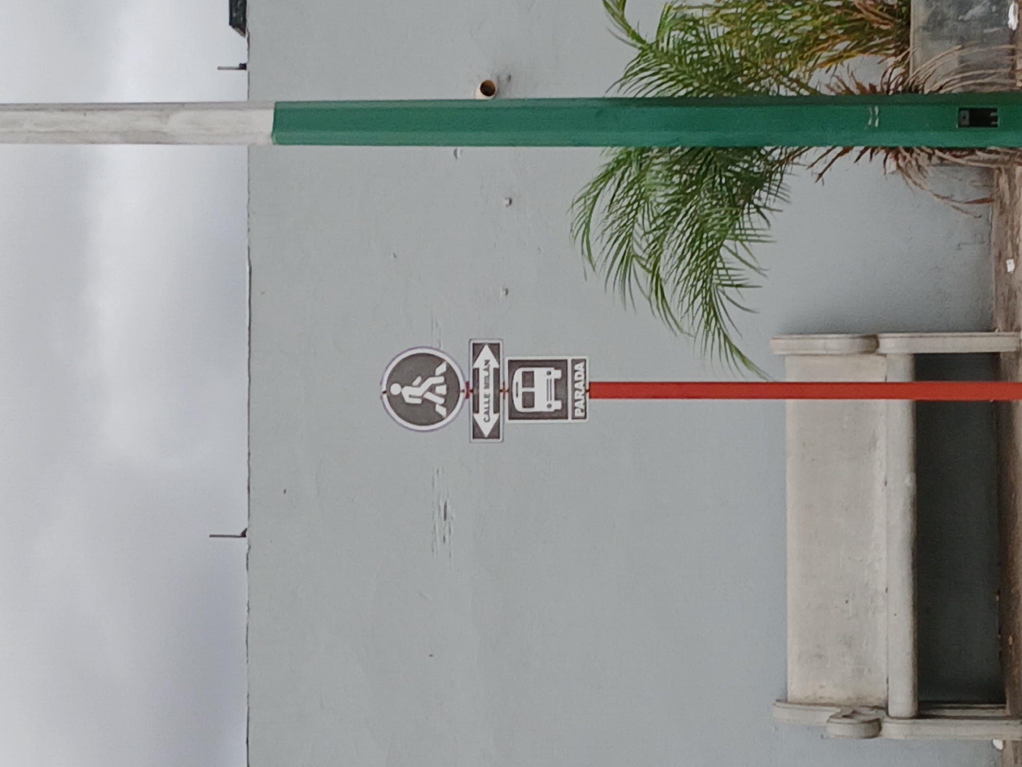 The image shows a sign on a wall in Ciudad Guayana, Venezuela. The sign indicates a pedestrian crossing and a bus stop, with the word "PARADA" (stop) visible. The wall is light-colored, and a green and silver bar is positioned horizontally above the sign. A palm tree is located to the right of the sign, and part of a concrete bench can be seen below. The sky is overcast.