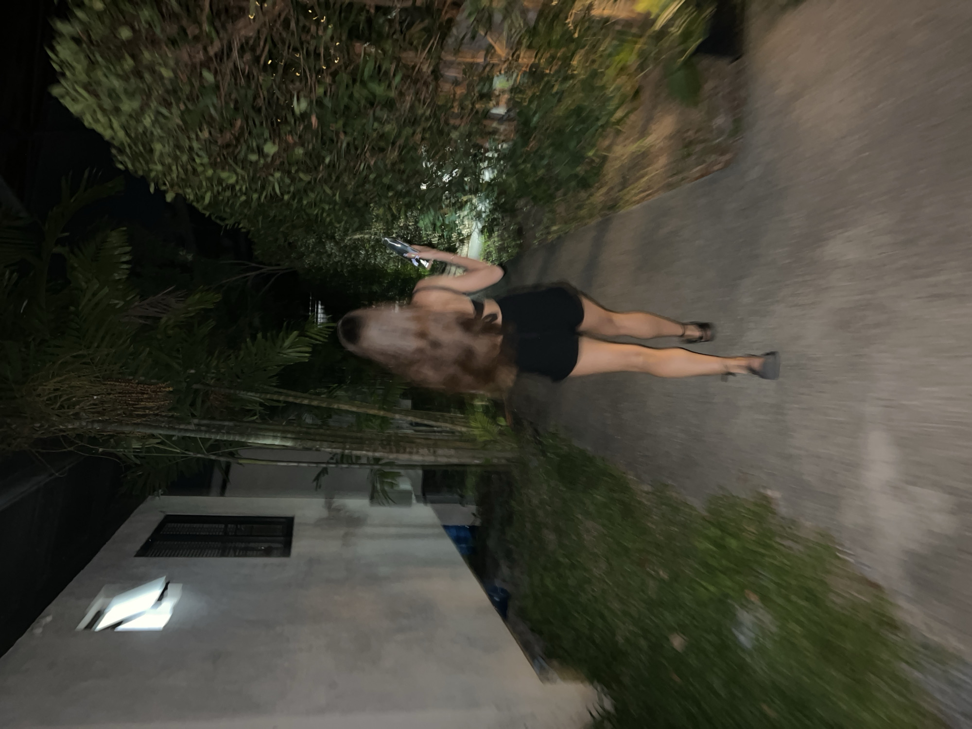 A blurred image depicts a person walking on a sidewalk in บ้านบางรักษ์, Thailand at night. The individual is female, wearing a black top and shorts, and is looking at a phone in her hand. The sidewalk is bordered by lush, green vegetation on both sides. A building is visible on the left. The scene suggests movement due to the blurry quality of the image.