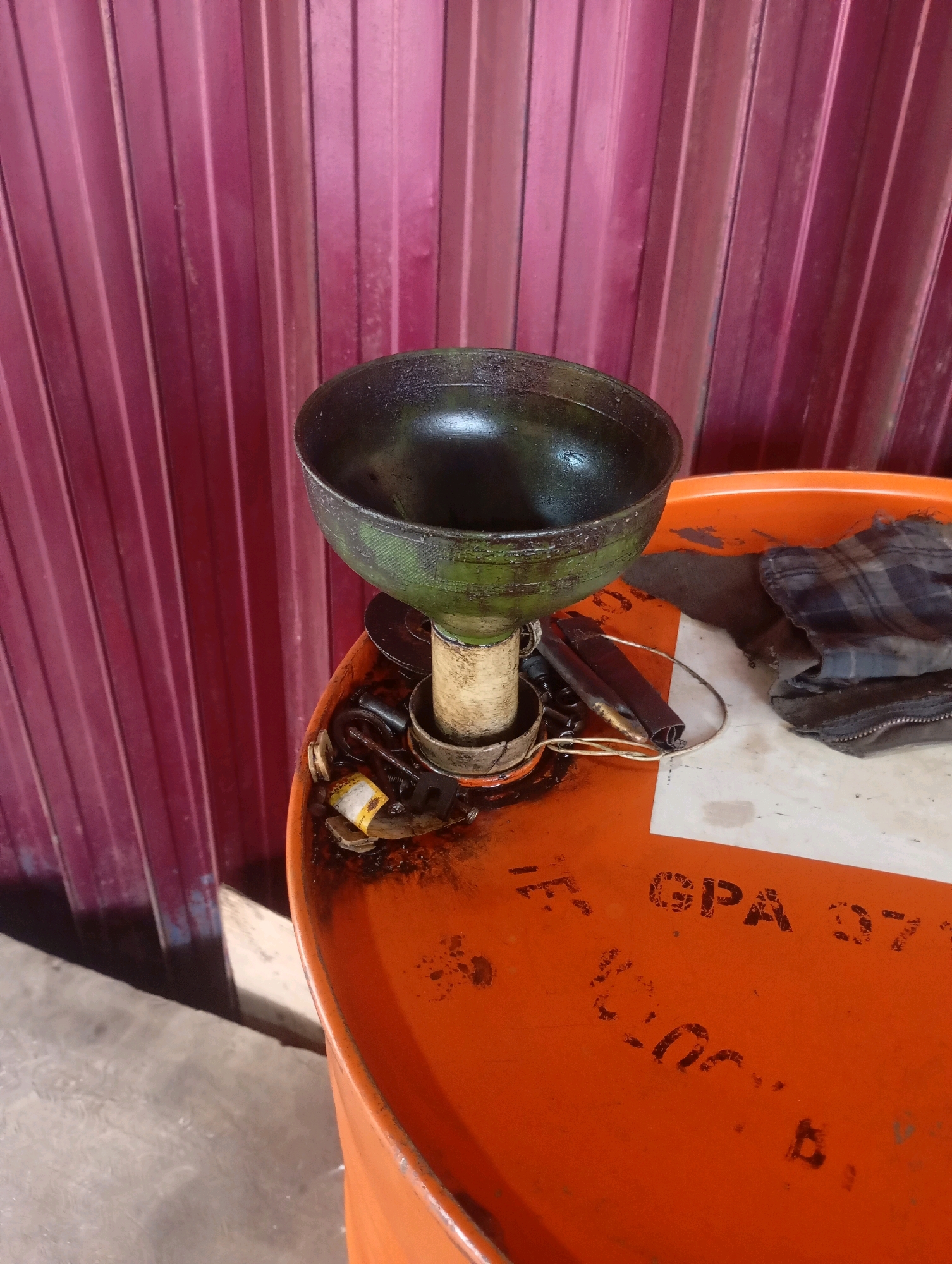 A metal funnel, with a green patina, is placed on top of a bright orange metal barrel. The barrel has some white markings on its surface, including "GPA 37". The funnel appears to be supported by a cylindrical object, possibly a pipe or a stand. Scattered around the base of the funnel on the barrel are several small objects, including what appear to be metal fasteners, a piece of yellow paper, and some dark fabric, possibly a bag or rag. The background consists of a vertical, ribbed, dark pink wall. The scene is indoors, with a concrete floor visible at the bottom of the image. The lighting suggests it may be daytime, with some shadows cast.