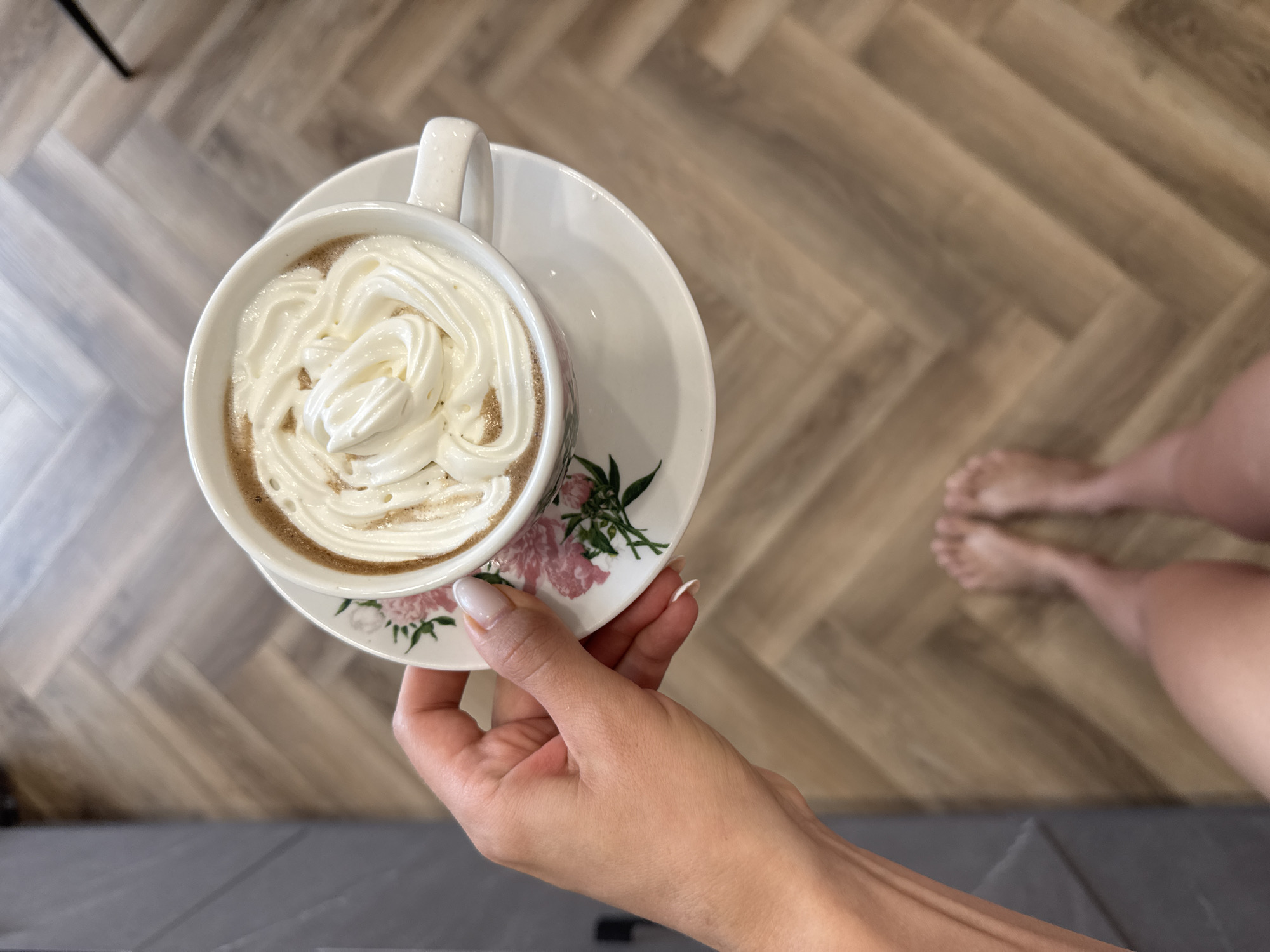 An overhead shot shows a person's left hand holding a white mug filled with a creamy beverage topped with whipped cream. The mug sits on a matching saucer decorated with a floral pattern. The background consists of a herringbone-patterned wooden floor. In the lower portion of the image, the person's bare feet are visible. The scene appears to be indoors, likely in a home, given the flooring and casual attire. The lighting suggests an indoor environment, possibly natural light.