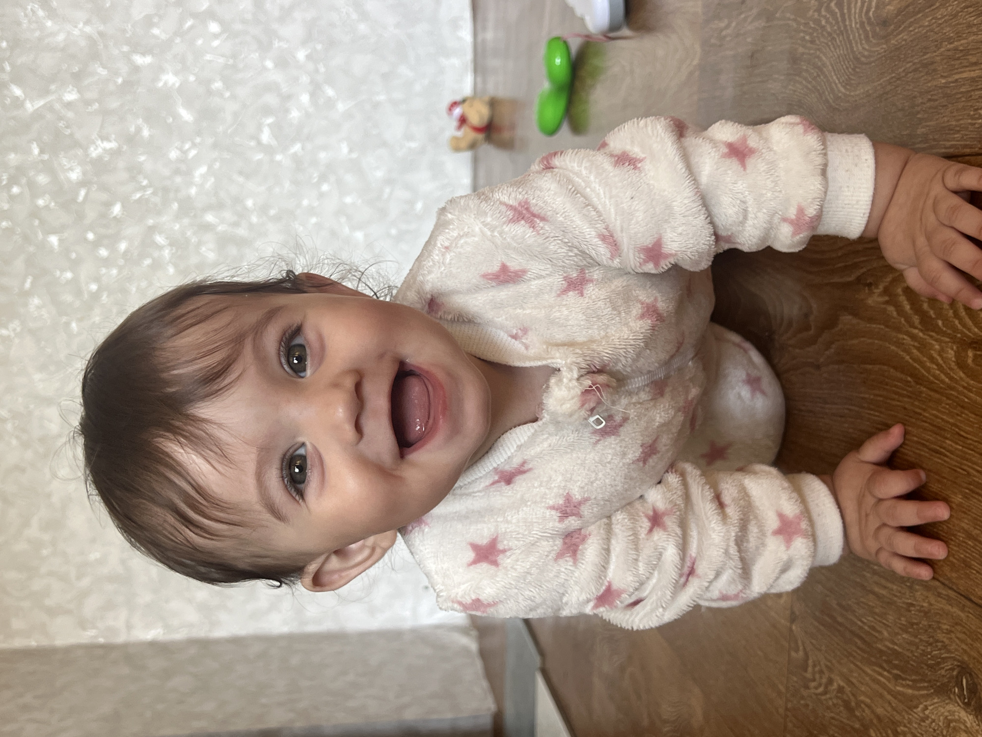 A baby lies on the floor, dressed in a white, star-patterned outfit, smiling with an open mouth. The floor is made of wooden panels, and the wall is white. Toys are scattered nearby. The location cannot be determined as Elena, Bulgaria, solely from the details in the image.