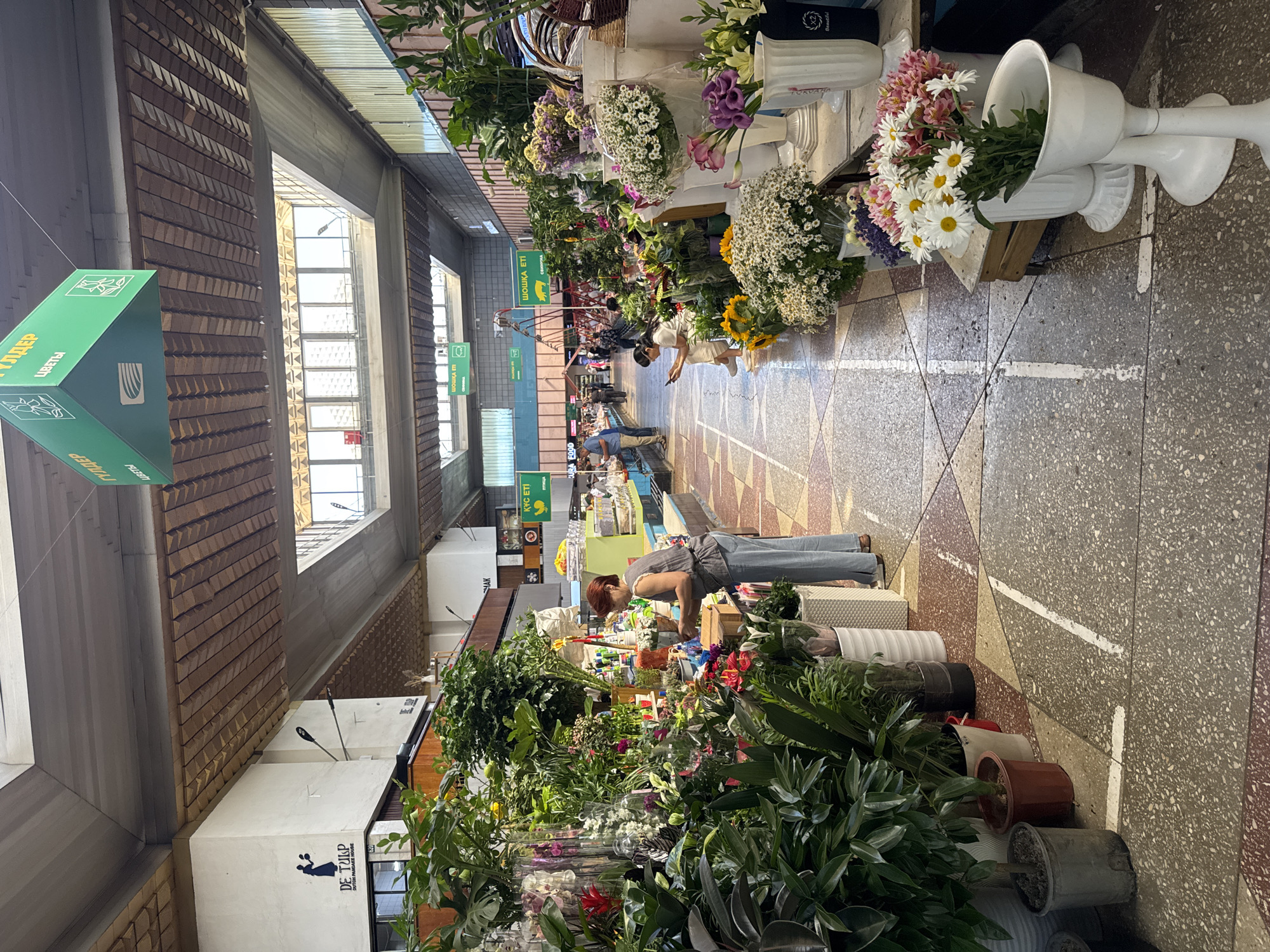 The image depicts a bustling indoor flower market, likely located in Almaty, Kazakhstan. The scene is characterized by rows of vibrant floral stalls and potted plants, with customers browsing and purchasing goods. Overhead, a unique architectural feature of the market's roof is visible, comprising wooden panels and skylights that allow natural light to filter into the space. Green signage, some with text in Cyrillic, hangs from the ceiling, indicating vendor names or categories.
In the foreground on the left, a woman with reddish-brown hair is tending to a large display of green plants and colorful flowers. She is wearing a grey sleeveless top and light blue trousers. Her posture suggests she is arranging or selecting flowers.
Further down the aisle, a woman in a white outfit is seated on a low bench, looking at her phone. She is surrounded by various potted plants and floral arrangements. Several other individuals are visible in the background, walking through the market or interacting with vendors.
The floor of the market is a mosaic of grey, red, and beige terrazzo tiles, with white lines delineating pathways. The stalls are laden with an abundant variety of flowers, including daisies, lilies, sunflowers, and various other blooms and greenery, arranged in vases, buckets, and pots. The overall impression is one of a lively and colorful marketplace.
