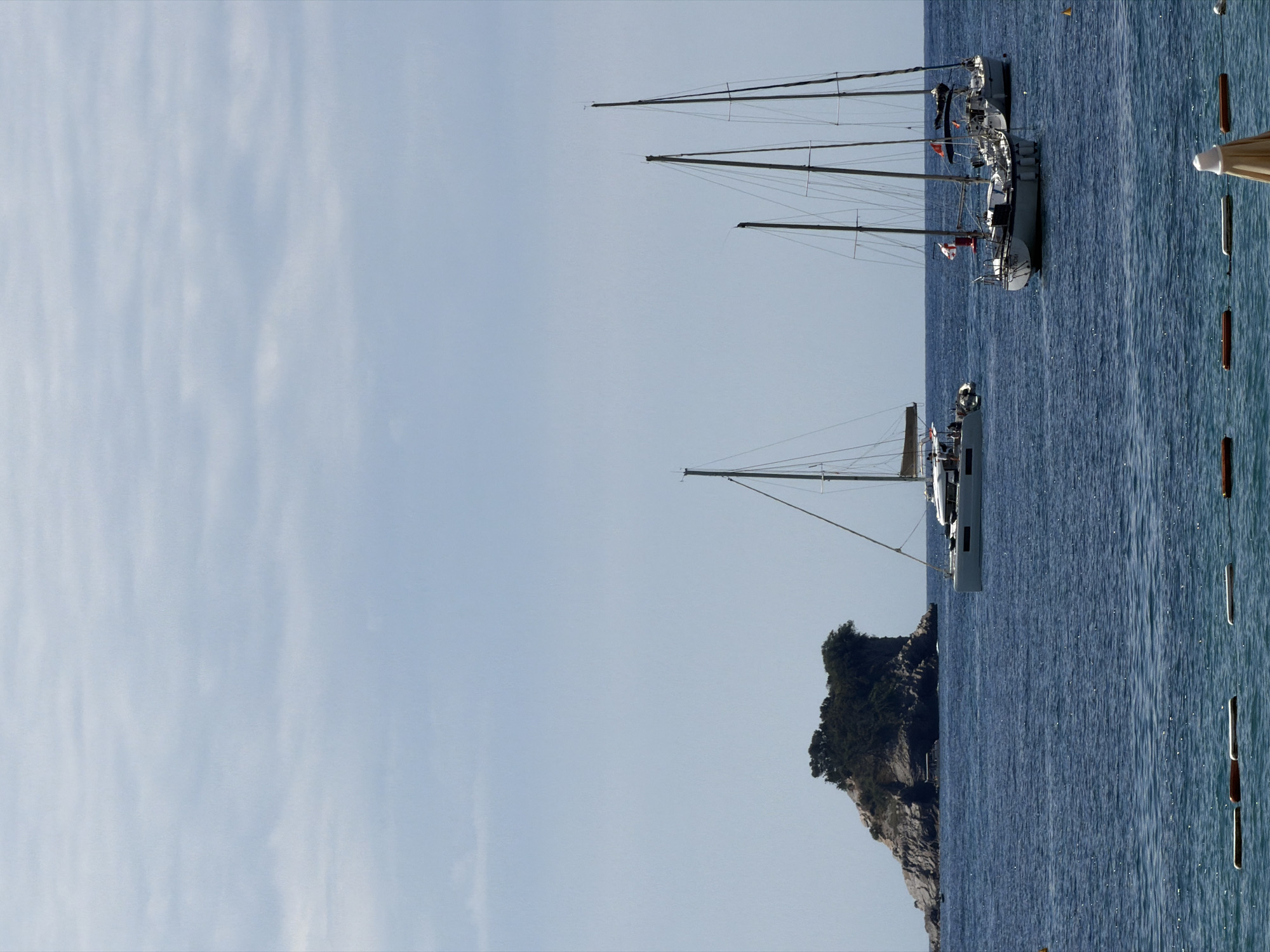 This image captures a serene coastal scene in Budva, Montenegro, featuring sailing yachts moored in calm, sparkling blue waters under a bright sky. The original image appears to be rotated, with the horizon running vertically. When correctly oriented, the upper half shows a pale blue sky with faint, wispy clouds, while the lower half displays the deep blue sea.
Several modern sailing yachts are anchored or moored in the water. In the mid-ground, a prominent white sailboat with its mast and rigging clearly visible floats to the right of the frame. A red flag and another smaller flag are flying from this boat. To its left, slightly further back, another sailboat is partially visible. The masts of at least two more boats extend into the sky on the far right, suggesting a popular mooring area.
On the left side of the frame, a rocky island or headland, densely covered with dark green vegetation, rises from the water, creating a natural backdrop. The water itself is calm, with sunlight reflecting off its surface, creating shimmering patterns. On the far right edge of the image, a series of vertical brown and white buoys or markers float in the water, possibly indicating a swimming zone or mooring points. A small part of a light-colored beach umbrella or similar structure is also visible on the extreme right.
The scene depicts a clear and peaceful day, likely morning or afternoon, given the bright light and tranquil conditions. No people are visibly present.