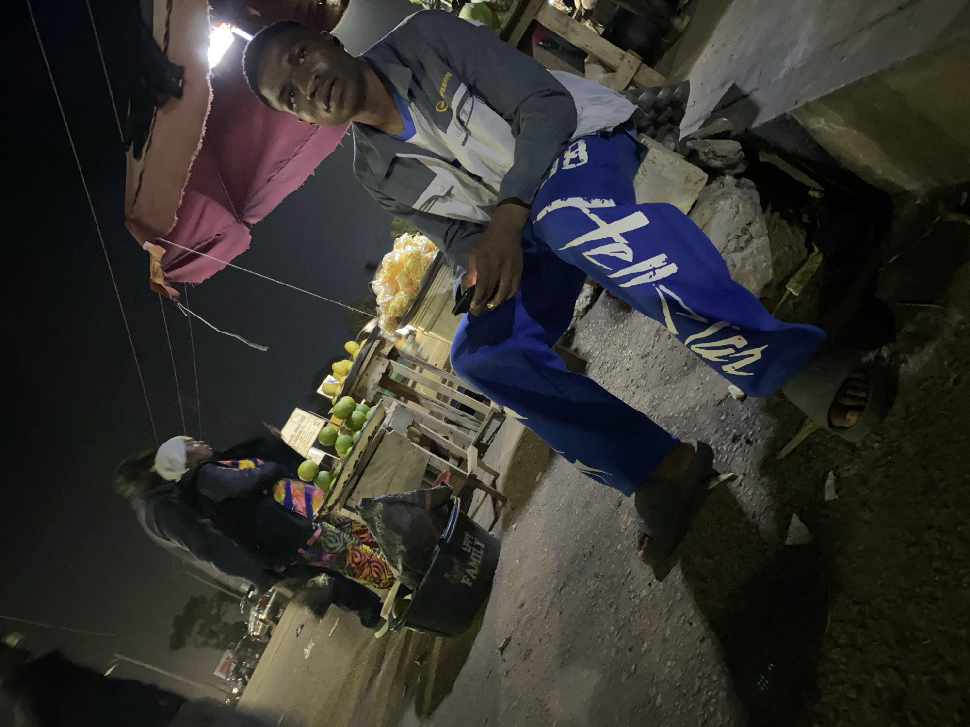 A medium shot shows a young Black man sitting on a pile of rocks at night, looking up and to his left. He is wearing a gray and white jacket over a blue shirt, and blue pants with white lettering. He holds a black mobile phone in his right hand. In the background, a woman wearing a patterned dress and headscarf stands near a fruit stand laden with green citrus fruits and yellow produce. A black plastic bucket with "APPI FAMILY" written on it sits on the ground to the left. The scene takes place outdoors on a dirt or asphalt surface, likely in a market area, with power lines visible overhead. The overall lighting suggests artificial light sources, creating shadows and a somewhat grainy appearance. The location is identified as Bukuru, Nigeria.