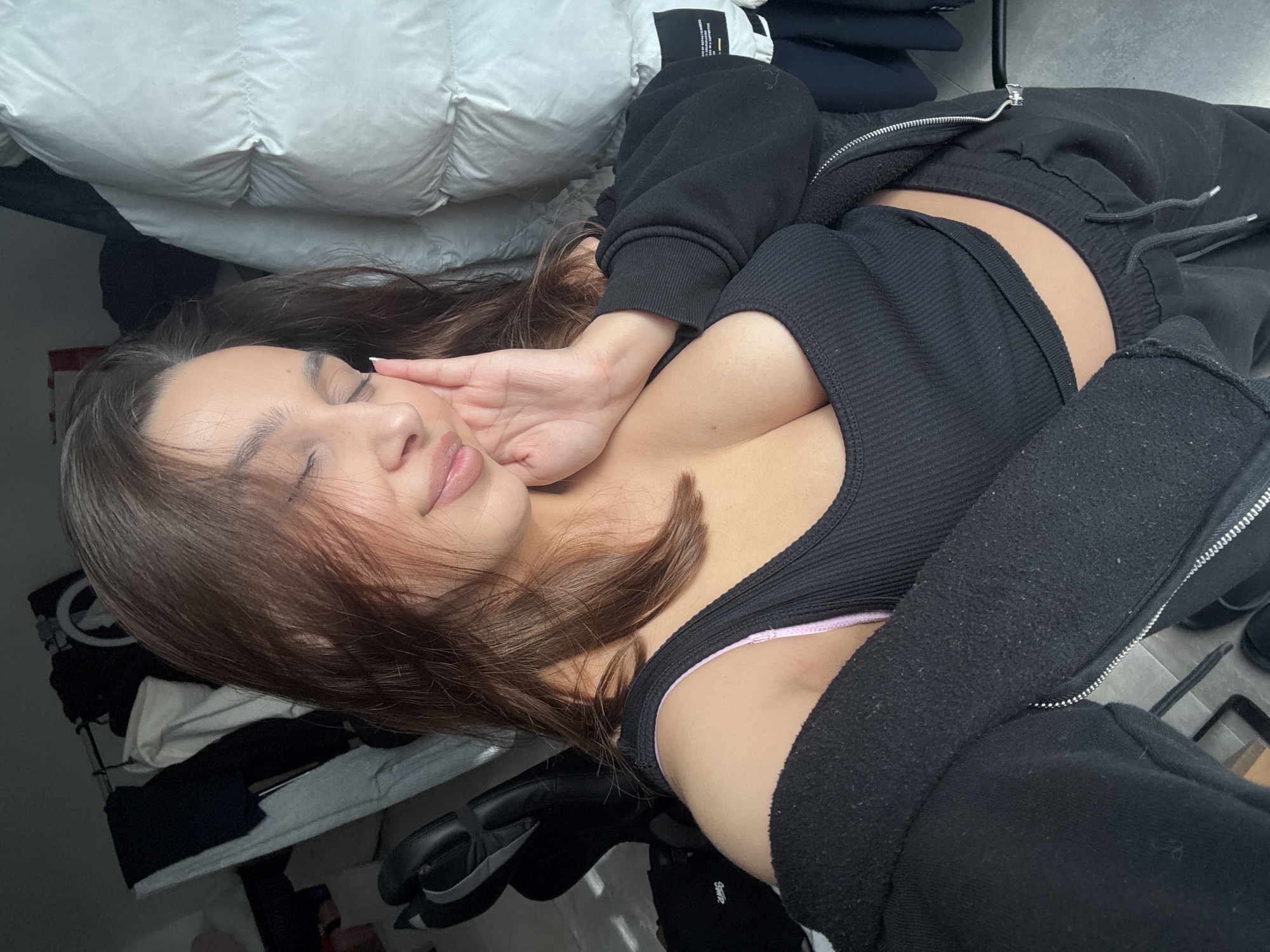 The image features a young woman lying down in an indoor setting, captured from a slightly high and tilted angle. She has long, dark brown hair and fair skin, with her eyes closed, suggesting a moment of rest or relaxation. Her lips are slightly parted. Her left hand is gently pressed against her cheek.
She is dressed in casual wear, including a black ribbed tank top, partially revealing a pink bra strap underneath. Over the tank top, she wears an open, black zip-up fleece hoodie, which shows some pilling. Black sweatpants with visible drawstrings complete her outfit.
The woman is positioned next to a large, white, quilted duvet or comforter, which dominates the upper part of the frame. The background shows a dark shelf or clothing rack with some folded garments. A pair of black headphones is visible on a surface in the lower right. The floor appears to be light-colored tile or concrete. The lighting is soft, indicative of natural daylight. A small, white product tag with indistinct black text is visible on a garment in the upper right background.