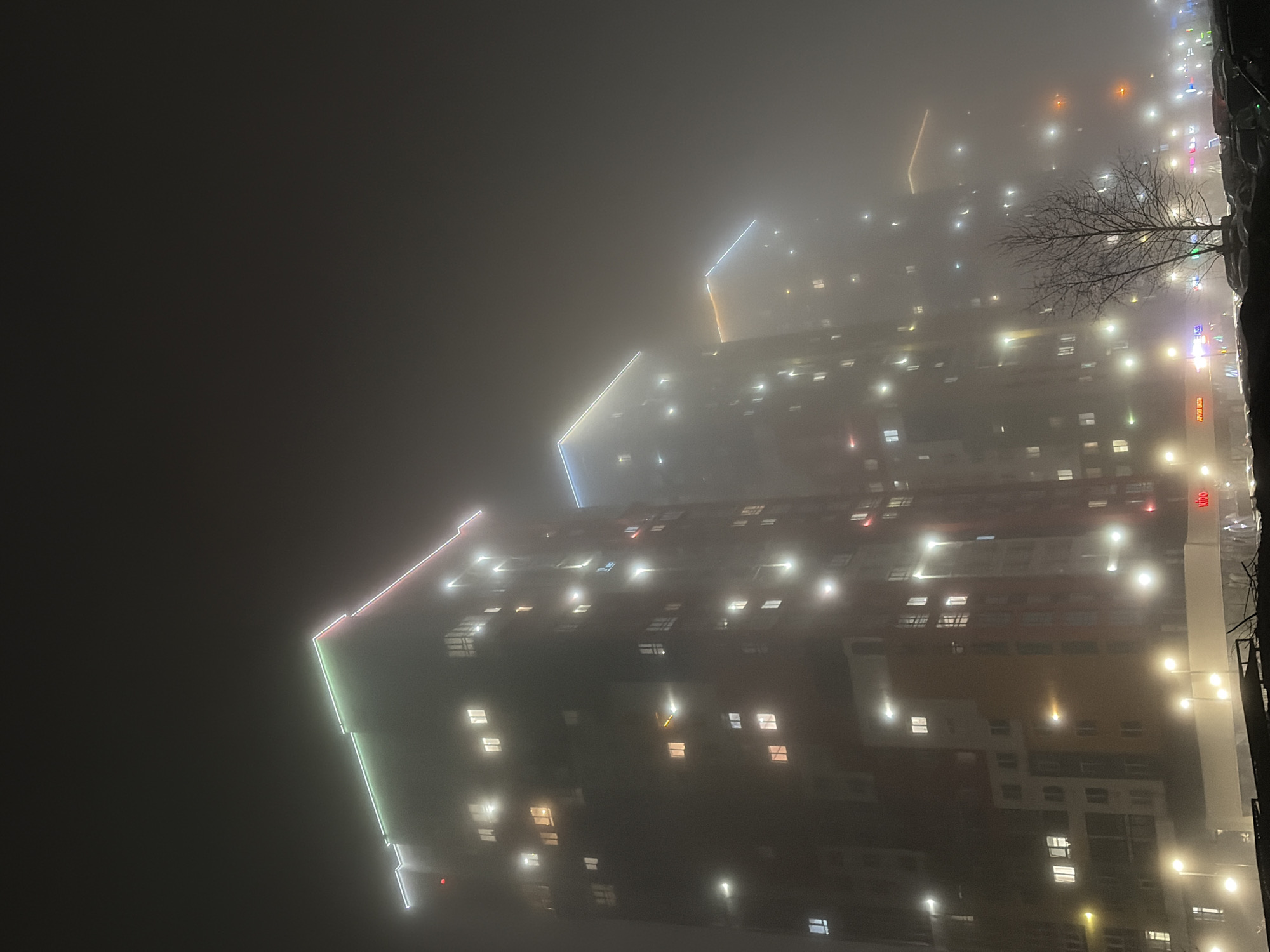 The image shows an illuminated residential building in Izhevsk, Russia at night in heavy fog. The building has distinct colorful neon outlines on its corners. Numerous windows are lit, indicating occupancy. The surrounding environment is obscured by fog, though street lights and other signs of urban life are visible. The image is taken from a relatively low angle.