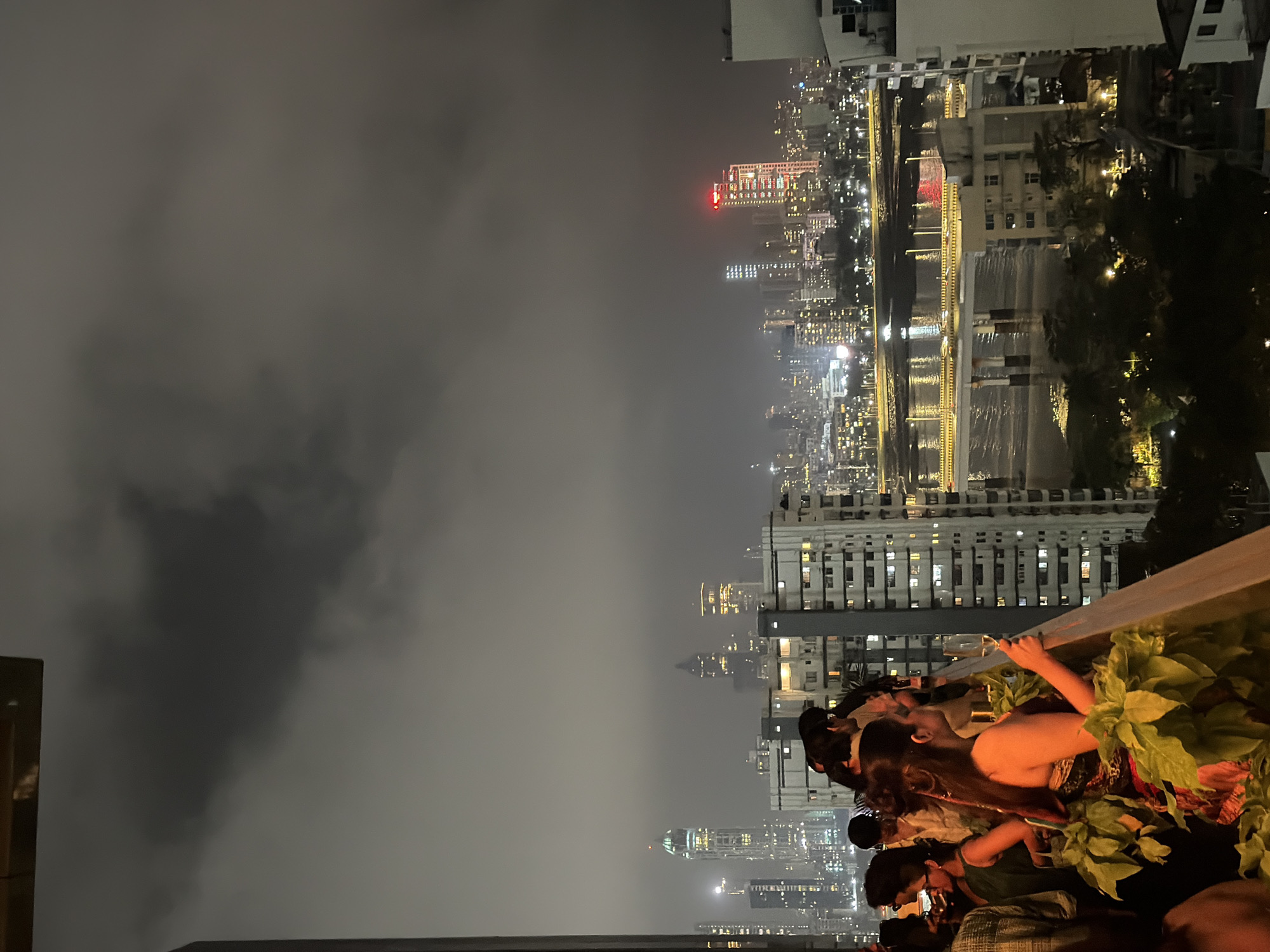 The image captures a nighttime cityscape of Mumbai, India, viewed from a high vantage point, likely a rooftop or balcony. In the foreground, a group of people are gathered, enjoying an evening on what appears to be a terrace or balcony. One woman in the foreground, wearing a colorful strapless dress, holds a glass of champagne and looks out at the city. Several other individuals are visible, some chatting, others looking towards the view. Lush green plants are present in the foreground, adding a touch of nature to the urban setting.
The background showcases the vibrant Mumbai skyline, illuminated by countless lights. Tall skyscrapers dominate the view, their windows glowing, creating a dazzling spectacle. A bridge, possibly the Bandra-Worli Sea Link, spans across the water, with its lights reflecting on the surface. The overall atmosphere is one of a lively urban gathering against the backdrop of a dynamic city at night. The sky is overcast with dark clouds, typical of Mumbai's weather, adding a dramatic effect to the scene.