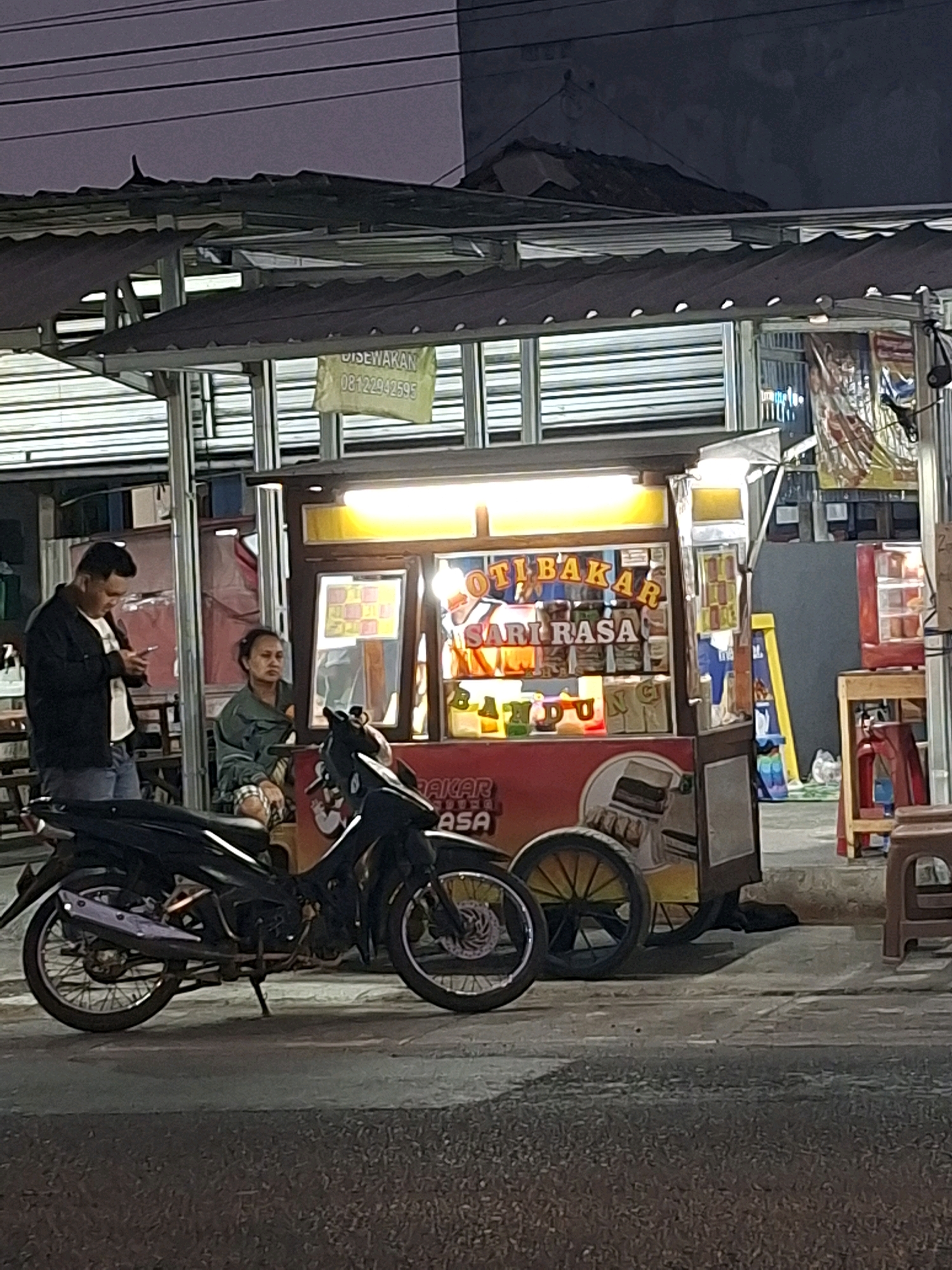 In Sidoarum, Indonesia, at dusk, a street scene unfolds with a food cart illuminated by bright yellow lights, displaying "ROTI BAKAR" (grilled bread) and "SARI RASA" (essence of flavor) in large, colorful letters. A man in a black jacket and jeans stands to the left, looking at his mobile phone. Beside him, a woman in a green jacket and patterned skirt sits on a chair. A black motorcycle is parked in front of the food cart. The environment appears to be an outdoor marketplace with a covered structure overhead. A sign with "DISEWAKAN 08122942595" is visible on the cart's awning.