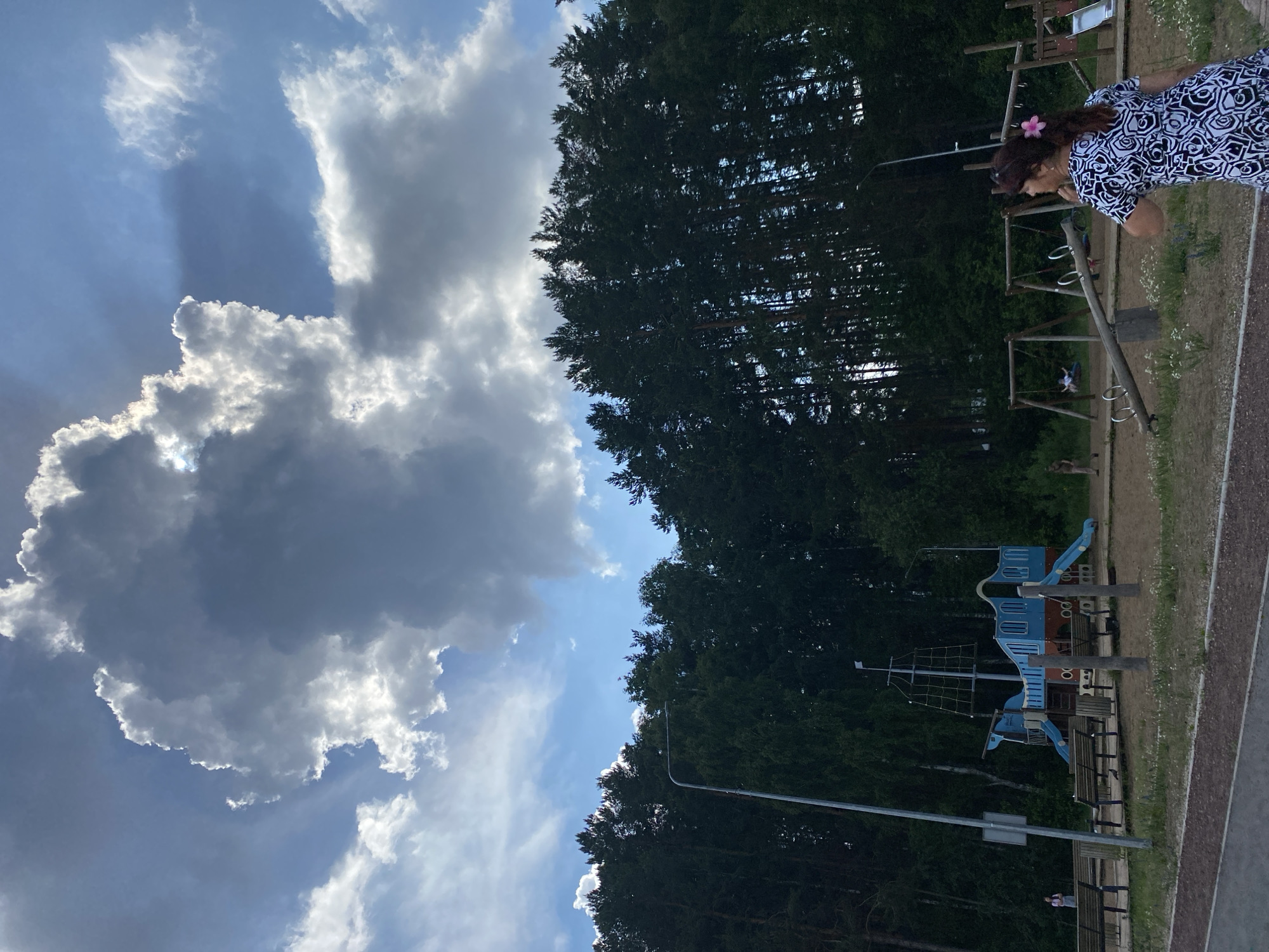 In the city of Chaikovsky, Russia, an outdoor scene unfolds under a dynamic sky. A prominent, large cumulus cloud dominates the upper portion of the frame, brilliantly illuminated by sunlight from the left, casting dramatic shadows. Below this celestial display, a dense line of dark green coniferous trees forms a natural backdrop. In the foreground, a public park area is visible. This park features a playground with a ship-themed structure, complete with slides and climbing nets, and several swings. Two women are present: one in the middle distance, standing near the ship playground, and another in the foreground, facing away from the camera and towards the playground, wearing a black and white patterned dress with a pink flower in her hair. The ground of the park is sandy, with scattered patches of green grass and small white wildflowers. A paved path runs along the bottom of the image, suggesting a recreational walkway. The overall atmosphere suggests a summer day in a park setting.