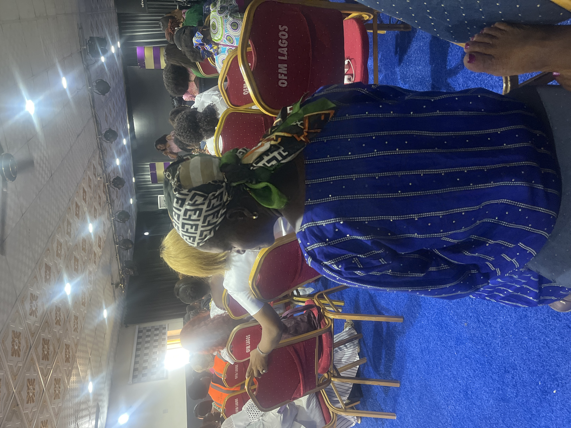 The image shows a gathering of people indoors, seated on red chairs labeled "OFM LAGOS." The foreground features a person wearing a blue striped garment and a head covering with a geometric pattern. In the background, there are additional attendees. The floor is blue, and a pattern on the walls indicates the interior of a building. The location is Ado, Nigeria.