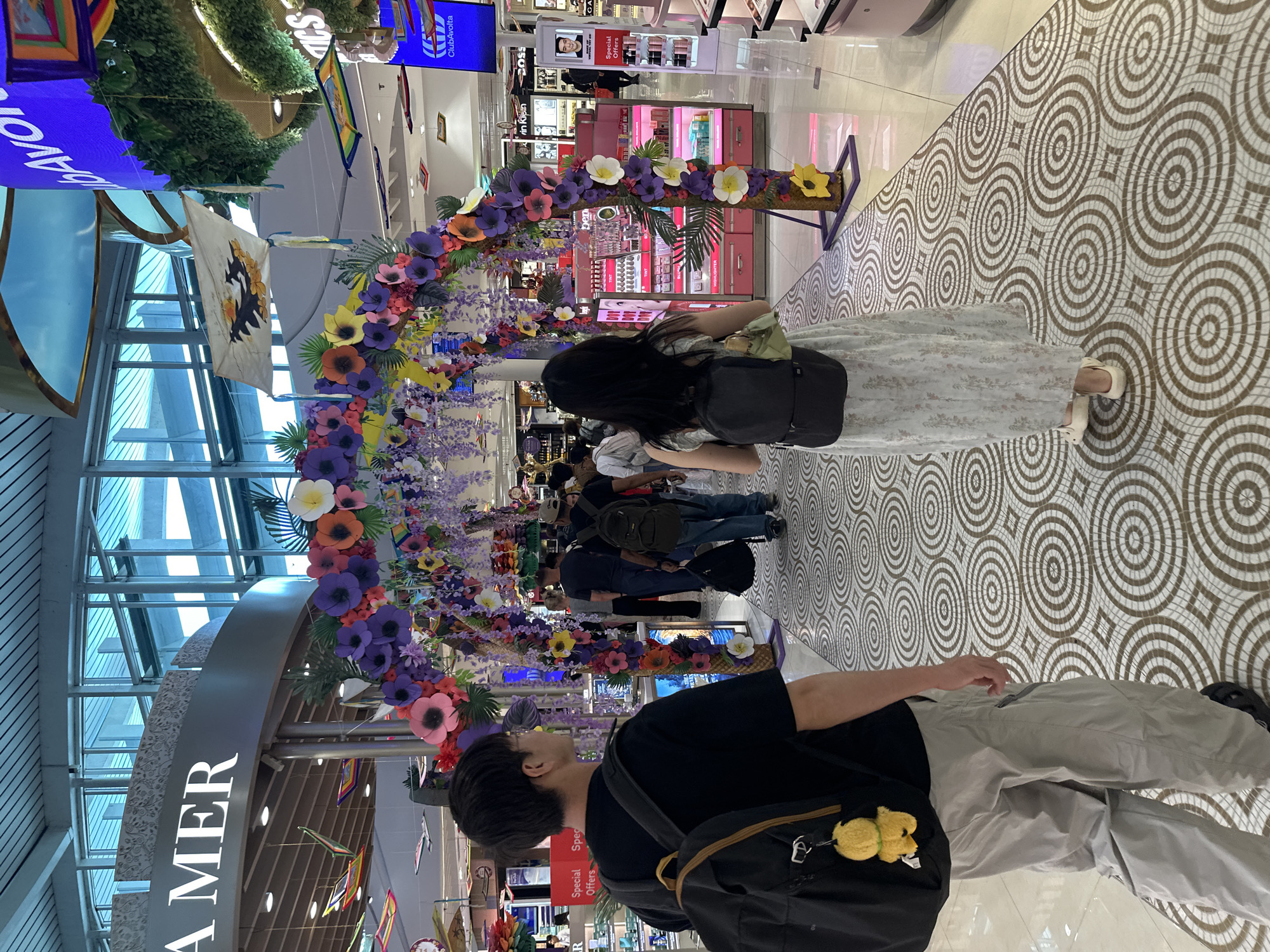 An overhead view shows a section of an indoor mall or airport terminal. A decorative archway adorned with colorful paper flowers and greenery is the central focus. The archway is positioned on a patterned tiled floor, creating a walkway for people. In the foreground, a man wearing a black t-shirt, grey pants, and a black backpack with a yellow charm is walking away from the camera, looking to his right. Behind him, a woman in a light-colored floral dress and a black backpack is walking through the archway. Other individuals are visible in the background, some walking and others browsing in shops. The shops appear to be retail establishments, with display shelves and signage. The ceiling features large windows and architectural elements. The overall scene suggests a public space with a festive or decorative theme.