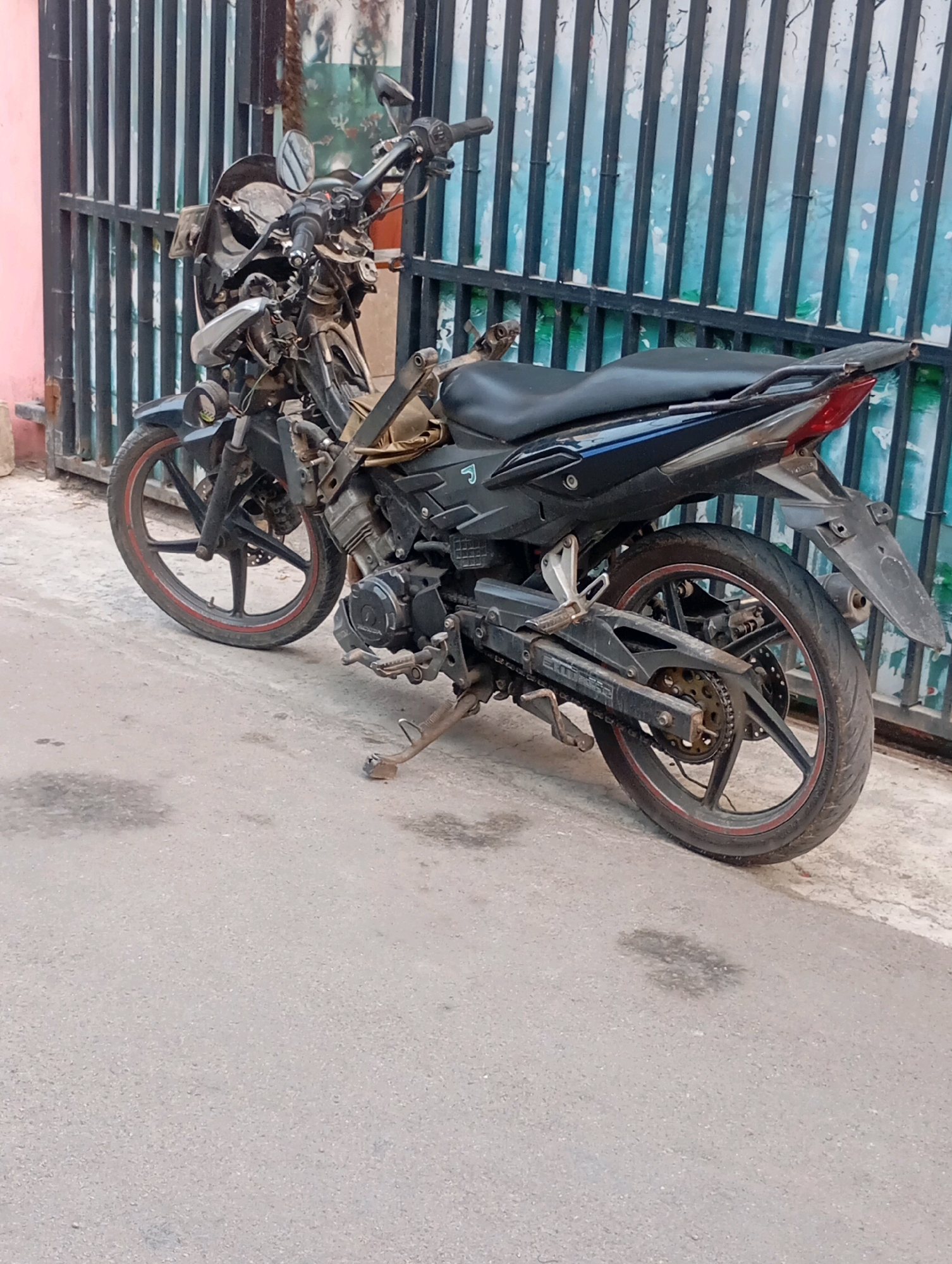 A dark-colored motorcycle with visible damage to its front fairing and handlebars is parked on a paved surface next to a black metal fence. The motorcycle appears to be a sportbike or a similar style, with black bodywork and red accents on the wheels. The front wheel is turned to the left, and the motorcycle is supported by its kickstand. The fence has vertical bars and is set against a wall with a blue and white patterned background. The ground is a rough, asphalt-like material with some dark stains, possibly oil or water. The scene suggests an outdoor environment in a city, consistent with a street or alleyway in Babelan, Indonesia. No individuals are present in the image.