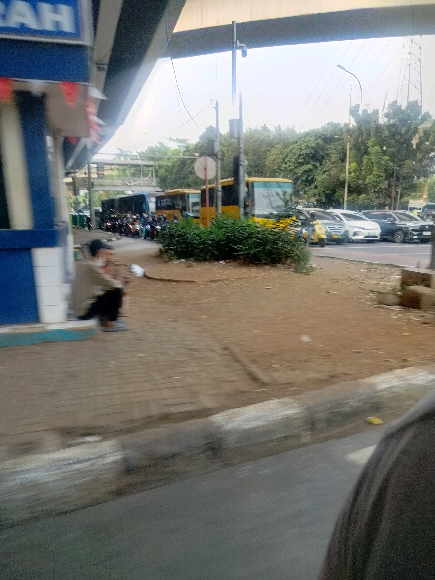 The image captures a street scene in East Jakarta, Indonesia, likely near a transit point under an elevated road. In the foreground, a person is crouched down on a paved surface, wearing a light-colored jacket and dark pants, with a dark baseball cap on their head. To the left, a blue and white structure, possibly a ticket booth or information center, is visible.
Further into the scene, several vehicles are present. A large yellow bus is parked, and behind it, a line of motorcycles and another bus, dark blue in color, are stopped. Beyond these, cars are parked and moving on the road. The environment includes trees and foliage bordering the road. Overhead, elements of an elevated road or bridge structure are prominent. The sky is bright, suggesting daylight, though the overall image is somewhat blurry and exhibits motion blur, indicating the photo was taken from a moving vehicle.
