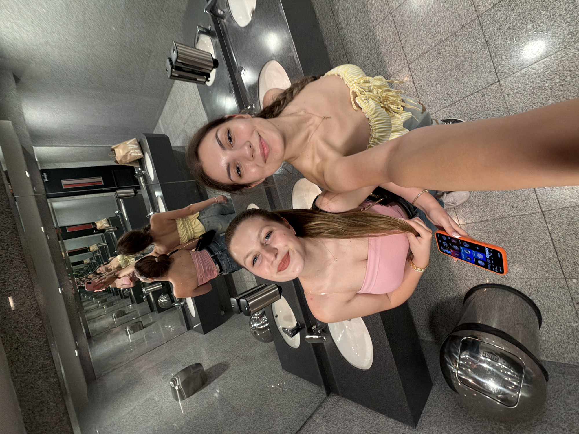 Two young women are captured in a selfie taken in a modern public restroom. The woman in the foreground, smiling at the camera, wears a pink strapless top and has light brown hair. Beside her, the woman holding the camera and taking the photo wears a yellow and white striped off-the-shoulder top and has darker hair. Both women appear cheerful and engaged.
The setting features a long, dark counter equipped with multiple white sinks and chrome faucets, along with stainless steel hand dryers or soap dispensers. A continuous mirrored wall behind the sinks creates an illusion of infinite reflections, showing the women and the restroom extending into the distance. The floor is a speckled grey tile, and the walls are a plain light grey. A silver trash can with a "PUSH" lid is visible on the floor near the woman in pink. In the background reflections, a brown paper bag can be seen on the counter.
The woman in the pink top holds a smartphone with a bright orange case, displaying various app icons on its screen. The image is taken from a slightly high angle, adding to the dynamic composition of the selfie. The overall atmosphere is casual and friendly, with the repeated reflections providing an interesting visual effect.