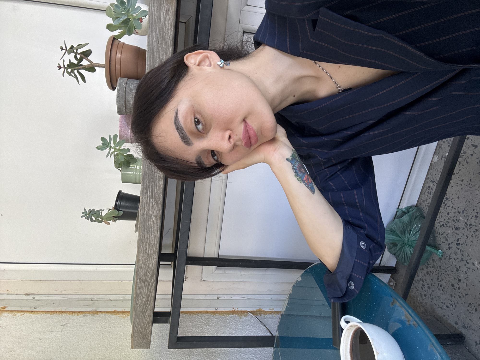 A young woman with dark hair and a butterfly tattoo on her arm is seated at an outdoor table, leaning on her hand with a pensive expression. She is wearing a navy blue pinstripe blazer and a silver necklace. A cup of coffee sits on the blue table beside her. Behind her, a shelf holds several potted plants. The scene is likely in Tbilisi, Georgia, though without further context, it is difficult to specify a precise location within the city. The background also shows the exterior wall and flooring material. A green tied bag is placed on the right part of the image.