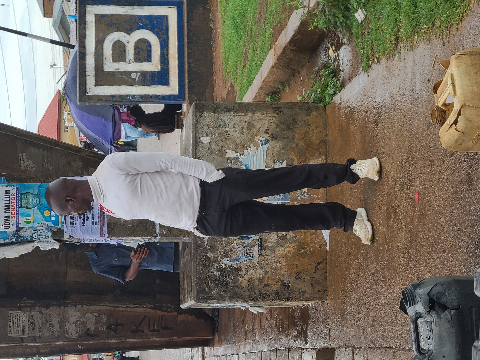 The image depicts a man standing in front of a concrete pillar covered with posters and advertisements in Gura Top, Nigeria. The pillar has political posters for Udya Mallum, a senatorial candidate, and other advertisements for insurance and employment opportunities. The man is wearing a white shirt and black pants, and he is standing on a wet, paved surface. In the background, there is a blue and white sign with a "B" on it, an umbrella, and some greenery. There are also damaged plastic containers visible on the ground. The overall environment appears to be a public space, possibly a street or marketplace.
