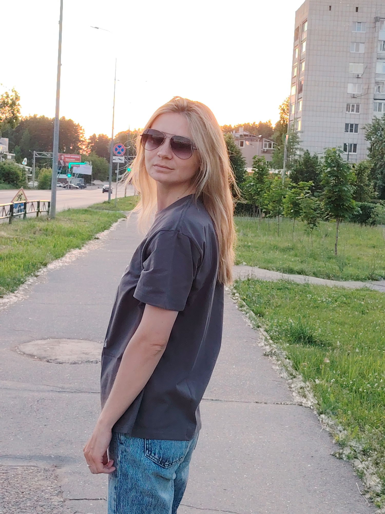 The scene is set in the city of Chaikovsky, Russia, depicting an urban or suburban street environment during late afternoon or early evening, indicated by the warm, golden light. A blonde woman, approximately 30-40 years old, with long, straight hair, stands on a concrete sidewalk, looking directly at the camera. She wears dark aviator sunglasses, a plain dark grey short-sleeved t-shirt, and faded blue denim jeans. A thin ring is visible on her left ring finger.
The sidewalk, featuring cracks and a circular manhole cover, runs diagonally, bordered on the right by a strip of green grass with small white flowers. Accumulations of white, fluffy seed material, consistent with poplar fluff, are visible along the sidewalk's edge.
To the left of the sidewalk is an asphalt road with visible lane markings. Several vehicles are present in the distance. Street infrastructure includes multiple lampposts, traffic lights displaying green, and various road signs, including a yellow pedestrian warning sign and a "No Parking" sign. Dense green foliage and trees line the road.
In the background, on the right, a multi-story, grey concrete apartment building with numerous windows and visible air conditioning units stands prominently. More trees and green space are visible around the building and in the mid-ground. The sky is bright, with a soft golden hue on the horizon, backlighting elements on the left side of the frame.