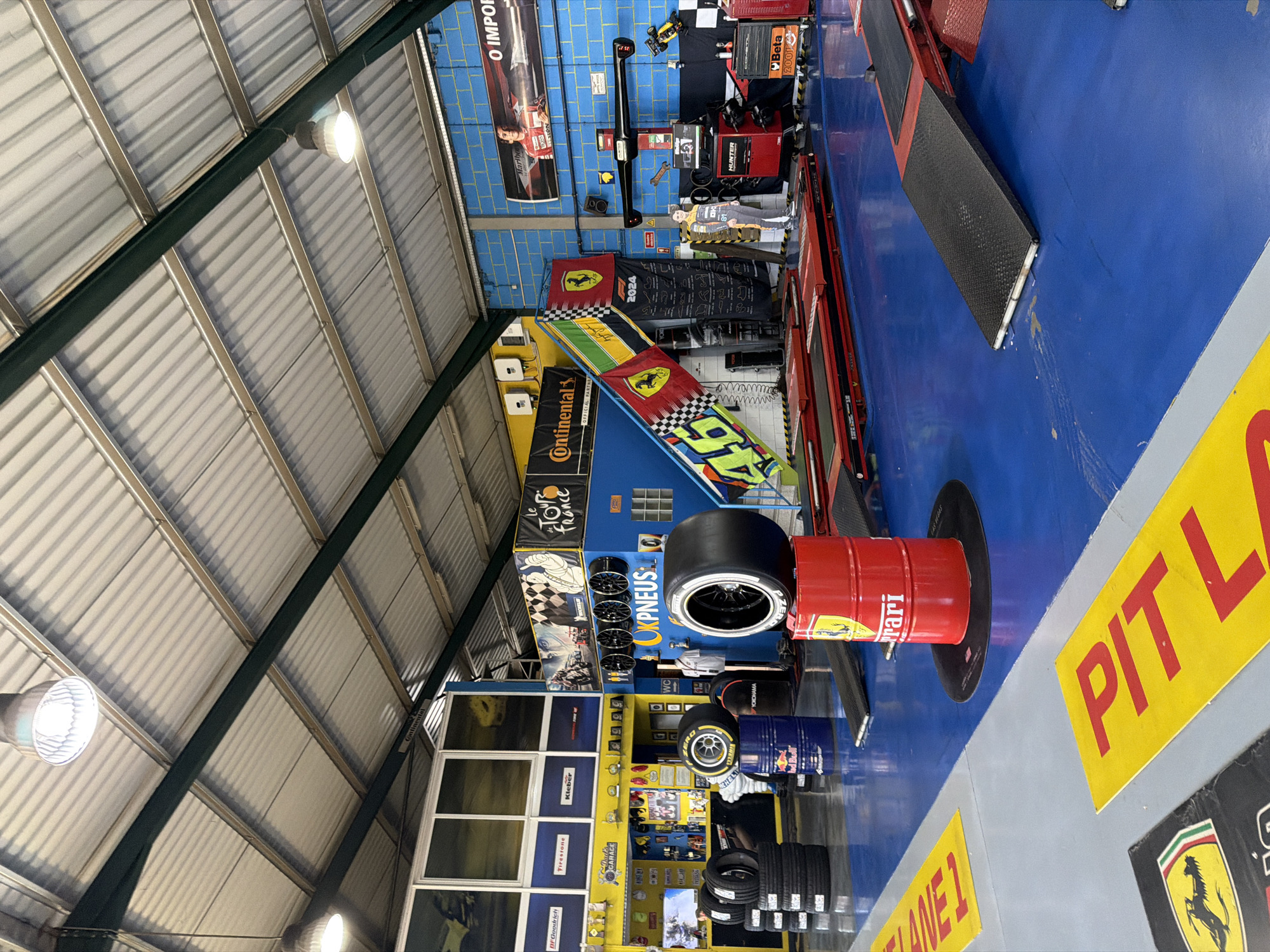 The image depicts the interior of an auto repair shop located in Vila Real, Portugal. The shop's ceiling is high and corrugated, illuminated by industrial lights. A blue and yellow color scheme dominates the walls, which are decorated with racing memorabilia including Continental tires, Pirelli tires, and racing flags. There is a variety of tires displayed throughout the shop. A red Ferrari-branded barrel is prominent in the foreground, next to a tire assembly. A “PIT LANE 1” sign is visible on the floor. In the background, various auto repair tools and equipment are present, including a Hunter wheel alignment machine and other tool cabinets. There is also a life-sized cutout of a race car driver in a racing suit. Several windows are also visible in the background.