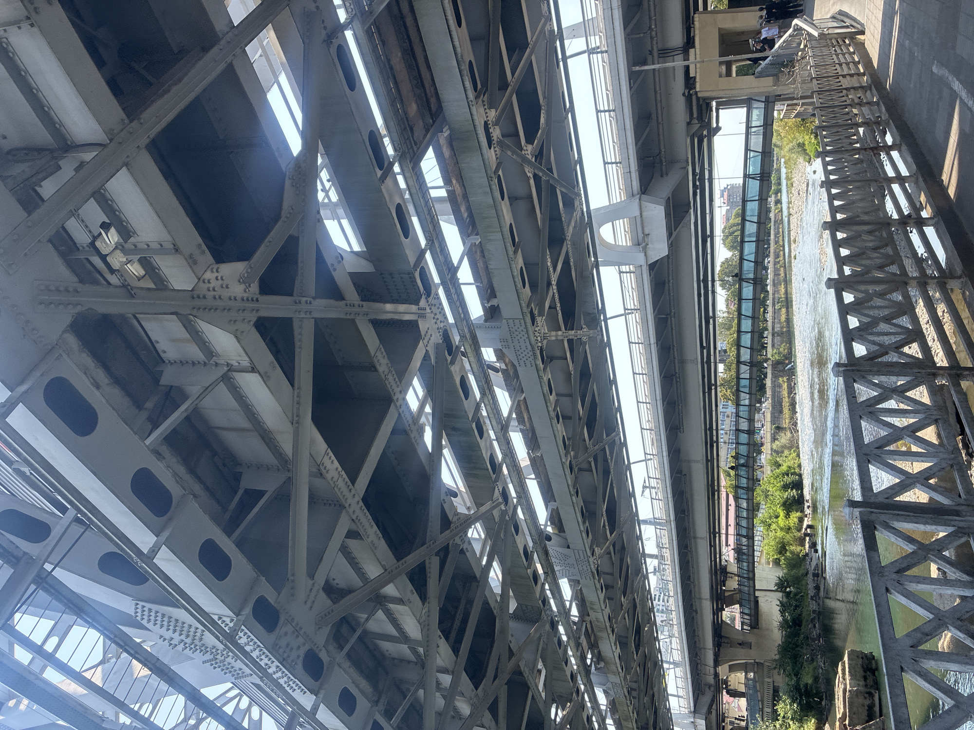 The image shows a view from beneath multiple bridges in Sochi, Russia. The primary focus is on the intricate metalwork of the bridge structures, with a clear sky visible through the gaps. Below these bridges, a river flows, bordered by green vegetation and rocky shores. A pedestrian walkway with a metal railing is in the foreground. In the distance, other bridges and buildings can be seen, indicating an urban environment. Several people are visible on the pedestrian walkway and on a bridge in the middle ground, some appearing to be dressed in formal attire. The scene suggests a sunny day with ample light illuminating the complex architecture.