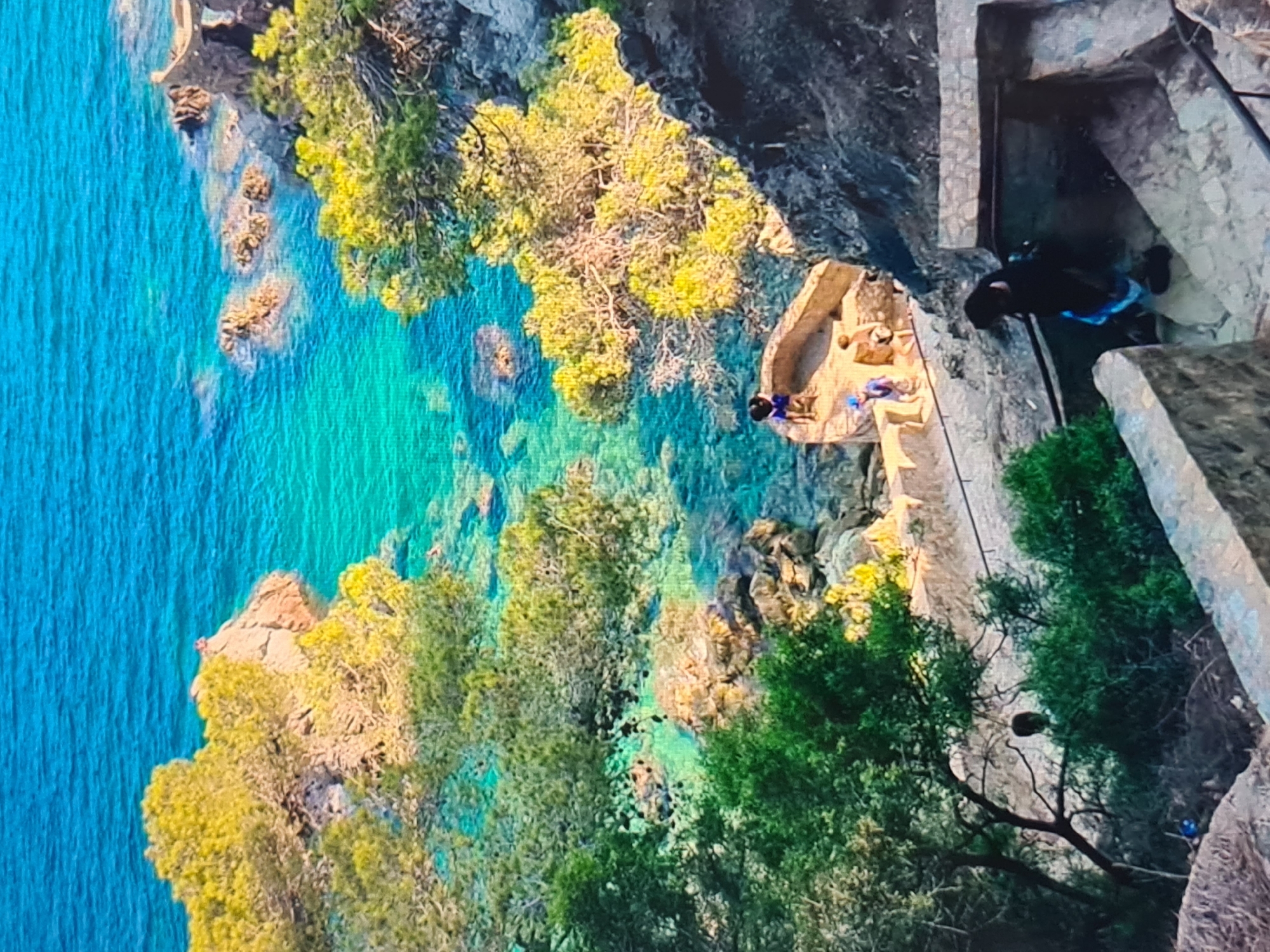 This image captures a vibrant, sunlit coastal scene from an elevated perspective, looking down a steep, rocky cliff face towards incredibly clear turquoise water.
The cliff is adorned with lush vegetation, primarily trees and shrubs with bright green and yellowish-green foliage, clinging to the rugged, dark gray and brown rock. Towards the center right of the image, man-made stone structures are integrated into the cliffside. A flat, light-colored platform or ledge extends from the cliff, offering a vantage point over the water. Two individuals are visible on this platform; one is sitting, dressed in blue shorts and a dark top, while another appears to be lounging nearby.
Adjacent to this platform, a set of stone stairs or a path is carved into the cliff, descending towards a shaded, recessed area that resembles a cave entrance or a built-in shelter. A third person, also wearing a dark top and blue shorts, is seen on these stairs, seemingly moving into or out of this shaded structure.
The water below is a striking feature, exhibiting remarkable clarity. It transitions from a deeper blue further from the shore to a brilliant, translucent turquoise closer to the cliff, where submerged rocks and the sandy bottom are clearly visible. Gentle ripples are present on the water's surface.
The scene depicts a beautiful, serene natural environment, likely a leisure or vacation spot, on a bright, sunny day. The strong sunlight creates clear shadows, emphasizing the textures of the rock and foliage. No visible text is present in the image.