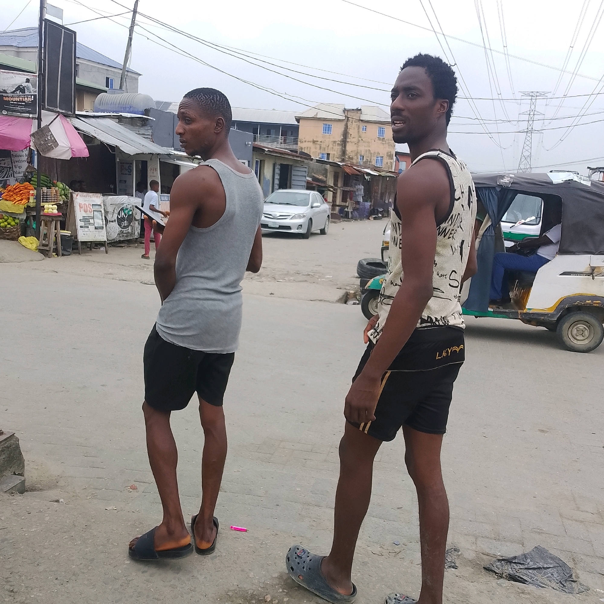 The image depicts a street scene in Ado, Nigeria, with two young men standing in the foreground, facing away from the camera. The man on the left wears a gray tank top and black shorts. The man on the right wears a patterned tank top with black shorts. They are standing on a paved road with some debris scattered on the ground. In the background, there are buildings, market stalls, a silver car, and a three-wheeled vehicle (tricycle). Power lines are visible crisscrossing the sky, and a large electricity pylon stands in the distance. The overall atmosphere appears to be that of a typical urban environment in Nigeria, likely a market area.