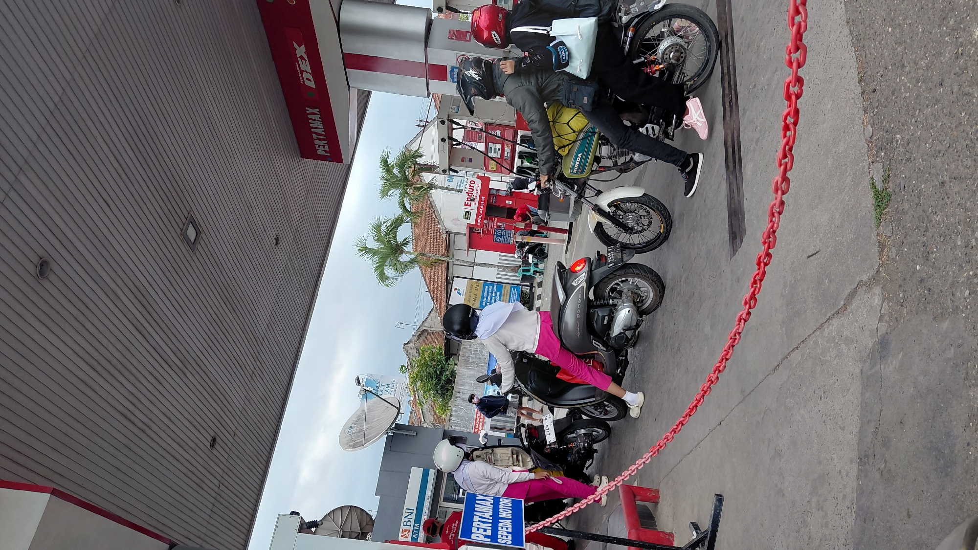The image shows a scene at a Pertamina gas station in Rejasari, Indonesia, where several people on motorcycles are waiting in line. A red chain barrier is visible in the foreground, separating the queue from the pumps. On the left, a person in a red uniform is attending to a customer on a scooter. Further back, two individuals are on scooters, one wearing pink pants and a white top, and the other on a dark grey scooter. In the middle ground, a person in a grey jacket is on a motorcycle, and another person in a red helmet is on the back. Palm trees and buildings are visible in the background, under a cloudy sky. The gas station features red and white signage, including "PERTAMAX DEX" and "SPBU 44.531.08". The ground is paved concrete.