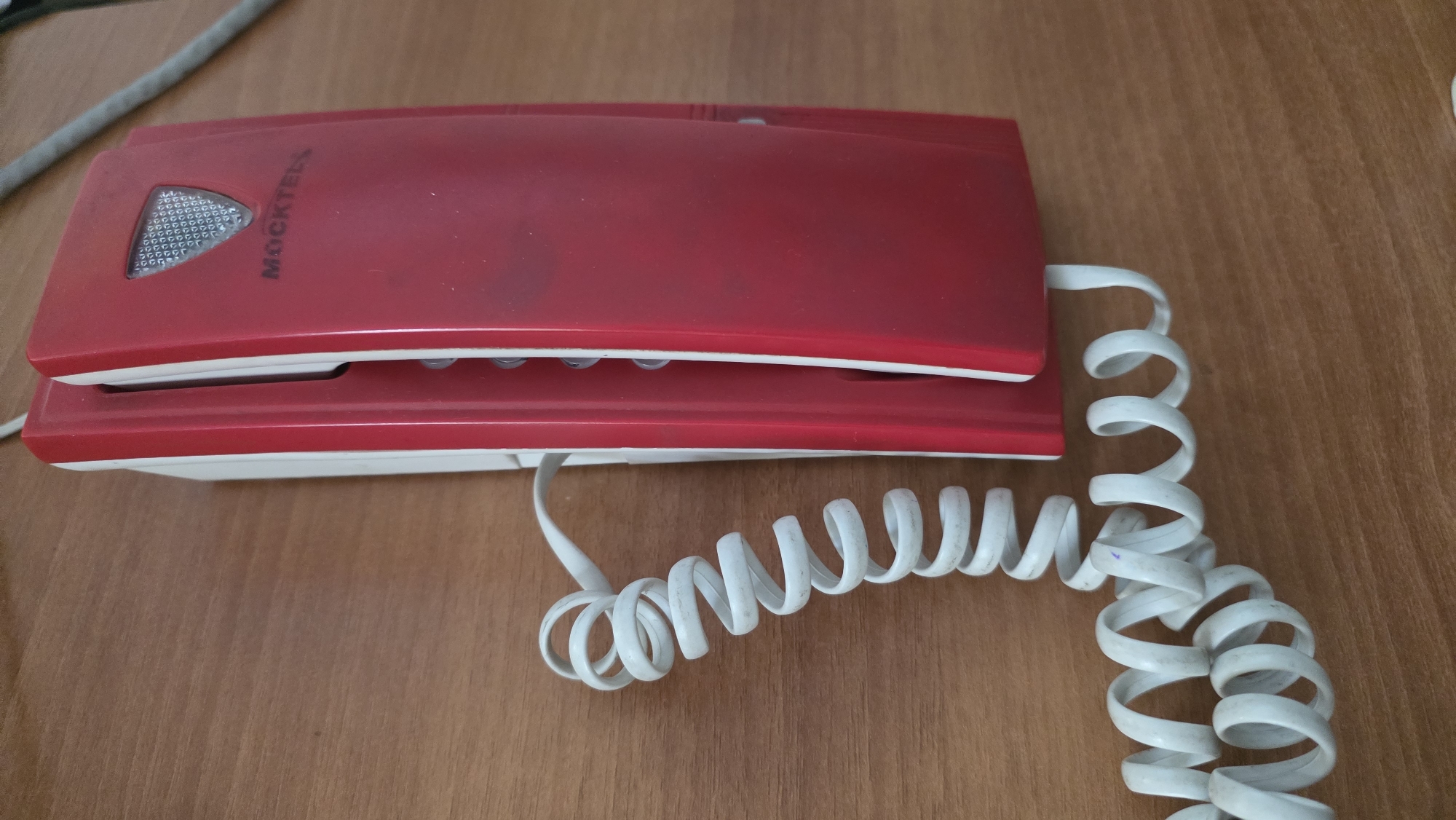 A red and white corded telephone rests on a wooden desk. The telephone has a red handset and a white base. A coiled white cord connects the handset to the base. The desk surface is made of light brown wood with visible grain. The telephone is positioned on the desk, with the coiled cord extending to the right of the frame.