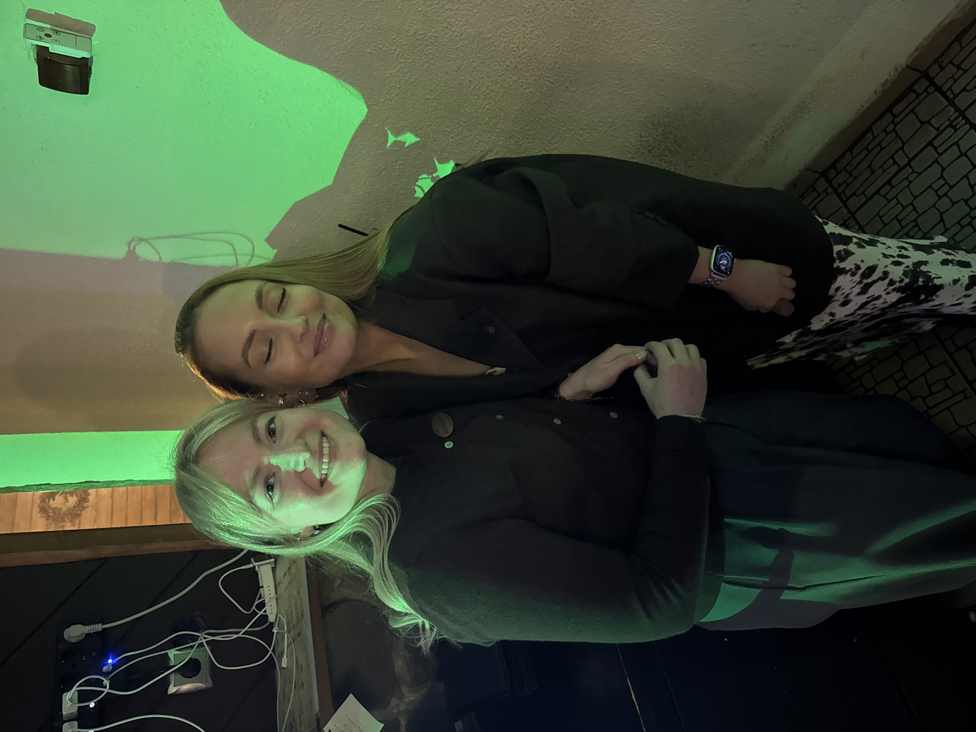 The image, oriented sideways with the left side of the frame as the top, displays two women posing together in an atmospheric indoor setting, likely a cafe or bar in Chaikovsky, Russia. The scene is dominated by a strong green light projected onto the walls and illuminating the subjects, suggesting an evening or nighttime ambiance.
In the foreground, a blonde woman smiles broadly, looking directly at the camera. She wears a dark, long-sleeved top, possibly a sweater or cardigan, and has her hands clasped. Behind her, a brunette woman with a subtle, contented smile and closed eyes rests her head close to the first woman. She is dressed in a dark jacket or blazer over a dark top, paired with distinctive black and white patterned trousers, resembling cow print. An Apple Watch with a metallic link band is visible on her left wrist.
The textured, light-colored walls in the background feature vibrant green projections, including small, silhouetted fish shapes, adding a whimsical touch to the venue's decor. In the upper left portion of the frame (when correctly oriented), an electronic device or light fixture with visible wiring is mounted on the wall. The lower left reveals a cluster of power cords, a white power strip with a glowing blue indicator light, and an electrical outlet against a dark wooden panel. The floor on the right side of the image appears to be a dark, textured surface, possibly tiles or cobblestones. A small, crumpled piece of paper rests on a dark surface in the bottom right corner, though no text is clearly legible. Both women exude happiness and relaxation, enjoying their time in the distinctively lit environment.