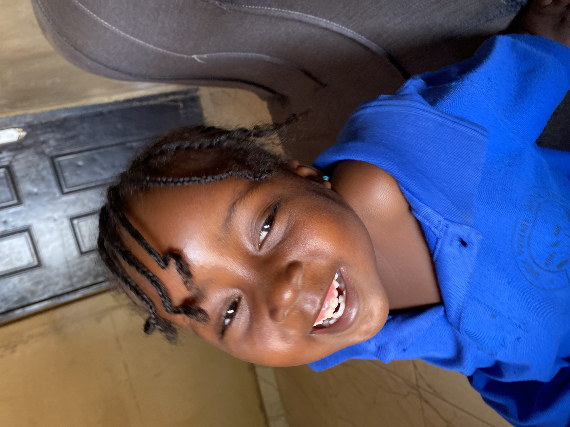 A young girl with dark skin and numerous small, dark braids is captured in a close-up, tilted portrait. She is wearing a vibrant blue short-sleeved t-shirt, which shows some signs of wear with a few small holes and marks. A small blue stud earring is visible in her right ear. The girl is smiling broadly, her eyes crinkling at the corners, and her mouth is open, revealing a mix of primary and possibly emerging permanent teeth. She looks directly at the camera with a happy and engaging expression.
The setting appears to be indoors in Kuru Dungus, Nigeria. In the foreground and background, a light brown or beige surface, possibly a wall or floor, is visible. Behind her head and shoulder, there's a dark, textured object, likely the back of a couch or chair. Further in the background, a dark, paneled object resembling a door or large piece of furniture can be seen. The lighting suggests a daytime setting. Faint, circular text or a logo is partially visible on the right side of her blue shirt, but it is illegible. The overall impression is a candid and joyful moment.