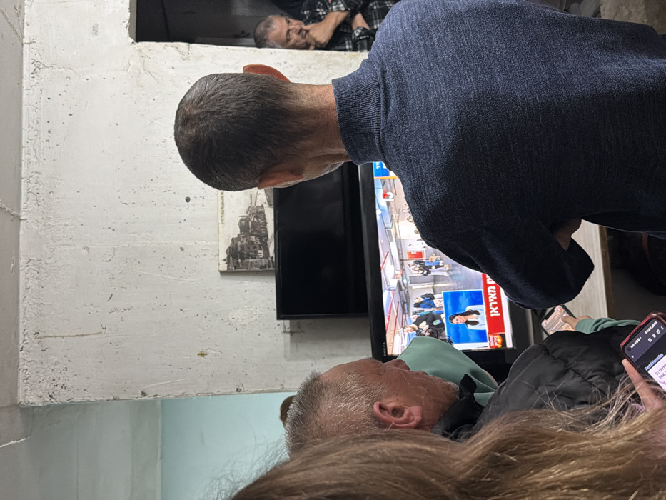 This image depicts an indoor scene in Tel-Aviv, Israel, where a group of individuals is gathered, observing a television news broadcast. The setting appears to be a utilitarian room with rough, light-colored concrete walls, one section exhibiting a pale blue-green hue. A flat-screen television, positioned on a dark stand, displays an Israeli news channel featuring a female anchor in the lower-middle of the screen, with a wider shot of an indoor public space (possibly an airport or public waiting area) above her. Hebrew text, including "מבזק" (news flash), is visible on the screen. Above the television, a small, dark-toned rectangular object and a black-and-white framed print depicting an old city scene are mounted on the wall. To the right, a dark doorway reveals a partial view of another space.
Four individuals are visible. In the immediate foreground to the left, the top of a person's head with long, light-brown hair is seen, alongside a hand holding a smartphone displaying Hebrew text. To their right, a middle-aged man with short, graying hair, wearing a dark jacket over a light garment, gazes towards the television. Central to the frame, a man with short, dark hair is seen from the back, wearing a dark blue turtleneck sweater, his arms appearing to be crossed. He is directly facing the television. In the background, partially obscured by the doorway to the right, another man with a beard and graying hair, dressed in a plaid shirt, looks towards the television with a contemplative expression, his hand near his chin. All individuals are oriented towards the television, indicating active observation of the broadcast. At least two individuals in the foreground are simultaneously engaged with smartphones.