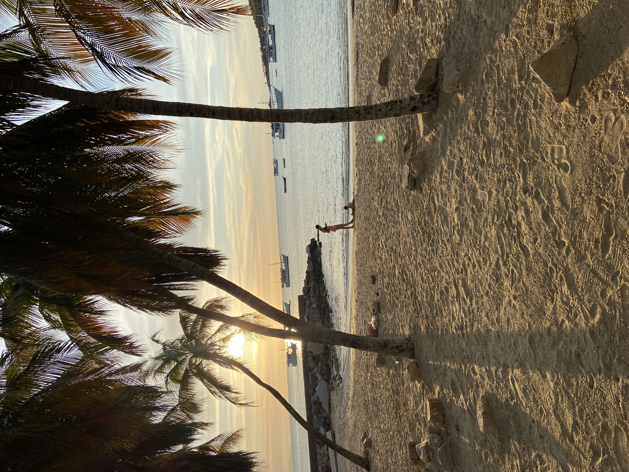 This is an outdoor, ground-level shot of a beach scene at sunset in Juan Griego, Venezuela. Palm trees frame the left side of the image and the top, with their fronds overlapping and partially obscuring the sky. The setting sun is visible between the trunks of the palm trees. The beach occupies the right side of the image, with sand showing footprints. In the distance, a line of rocks or a small wall separates the sand from the water. Two people are seen standing on the rocks, possibly interacting. Several boats are anchored in the water, which reflects the colors of the sunset. The overall atmosphere is warm and tranquil.