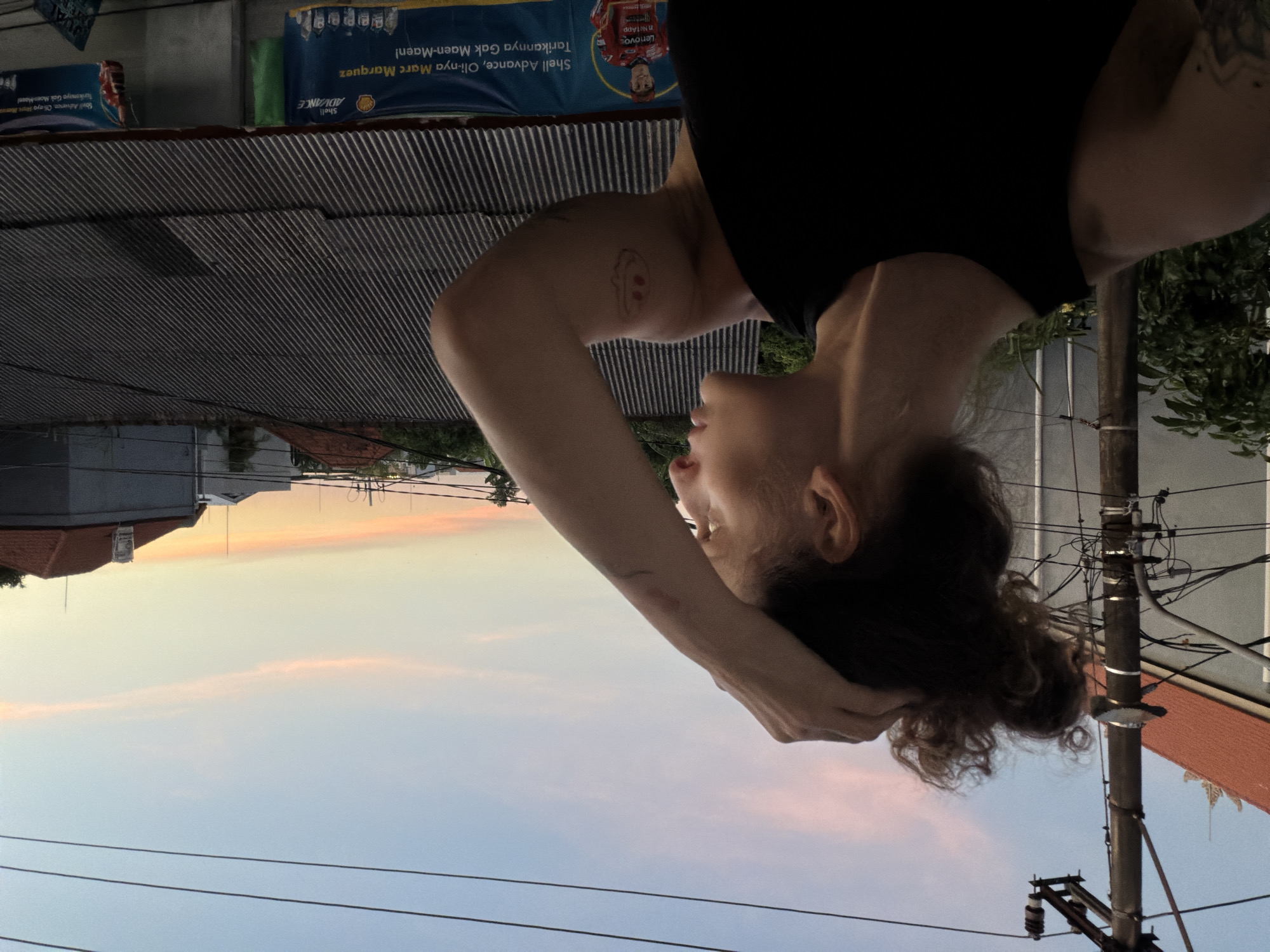 The image depicts a person, likely female with curly hair, wearing a black shirt, standing outdoors. The background shows a cityscape at dusk or dawn, with buildings, power lines, and a sky tinged with pink. There's a banner visible advertising Shell Advance motor oil with the slogan "Tarikannya Gak Maen-Maen!", suggesting a location in Indonesia, specifically near Kuta Utara (North Kuta), Indonesia, given the language and likely advertising. A metal corrugated roof is visible. The perspective of the shot is unusual, giving the impression that the person is positioned upside down.