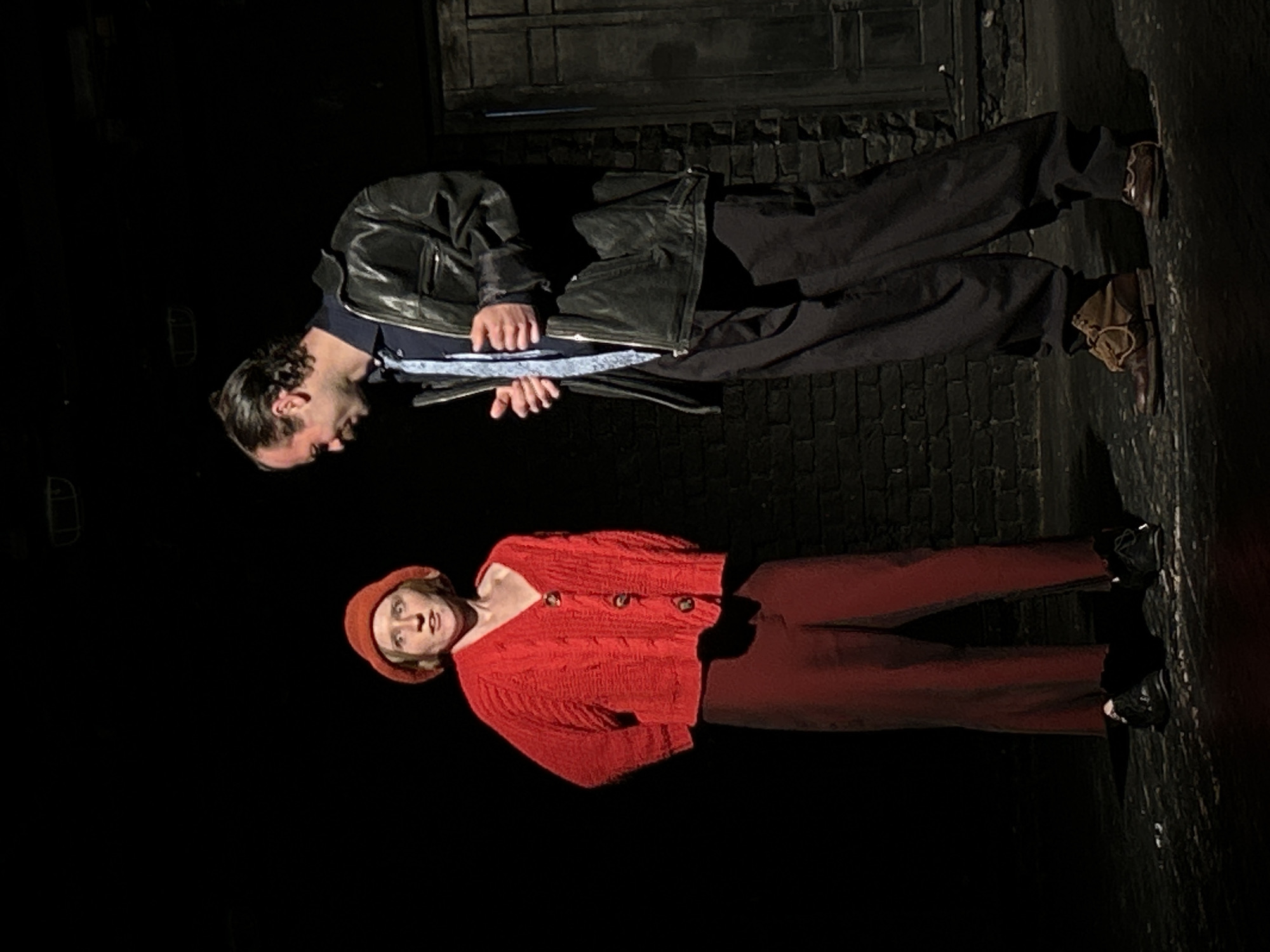 Two individuals are captured in a dimly lit environment, likely an urban setting. A male figure, dressed in dark pants, a black leather jacket, and brown shoes, is positioned in the upper part of the frame. He appears to be looking down, possibly holding something. Below him, a female figure is dressed in red clothing, including a cardigan and a hat. Her gaze is directed towards the camera. The background appears to be a brick wall or building facade, suggesting a city location of Unknown, Unknown.