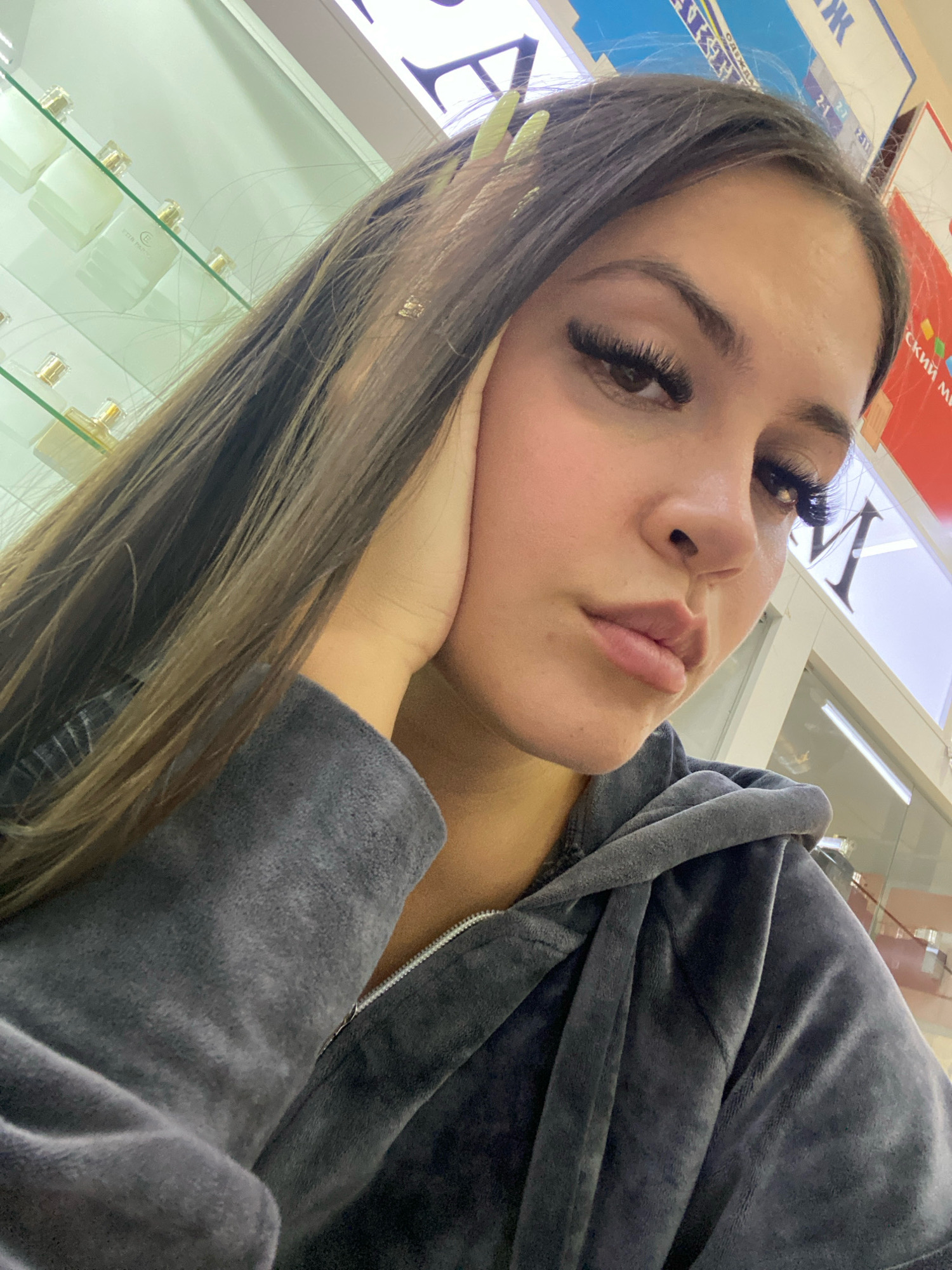 A young woman with long, dark brown hair and long, dark eyelashes is leaning her head on her hand, looking towards the camera. She is wearing a dark gray velour hooded jacket. The background is out of focus but appears to be a commercial establishment, possibly a store with glass shelves displaying bottles of perfume, and large advertisements or signs on the wall. The lighting is bright and appears to be artificial. The location is Chaikovsky, Russia.