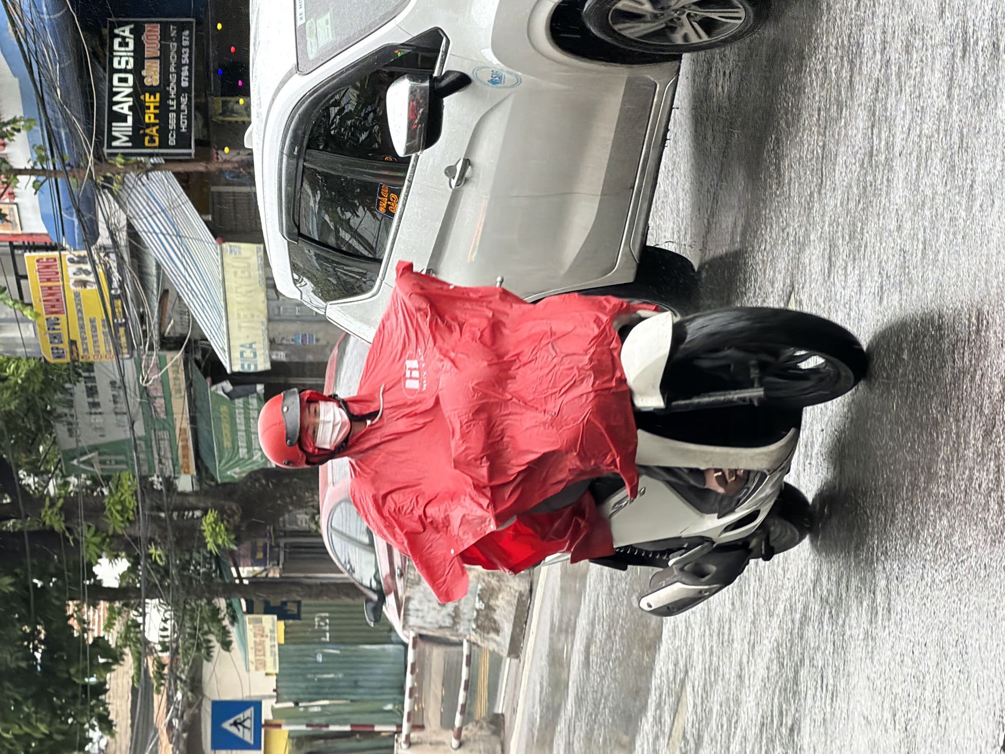 In Nha Trang, Vietnam, during a rain shower, a person on a white scooter wears a bright red poncho and a red helmet with a white face mask. They are riding on a wet, reflective street alongside a silver SUV. The SUV has a blue circular sticker on its rear door. In the background, various signs are visible, including one for "MILANO SICA CA PHE SON VUON." The street is lined with buildings and trees, with electrical wires overhead. A blue and white pedestrian crossing sign is present on the left side of the image.