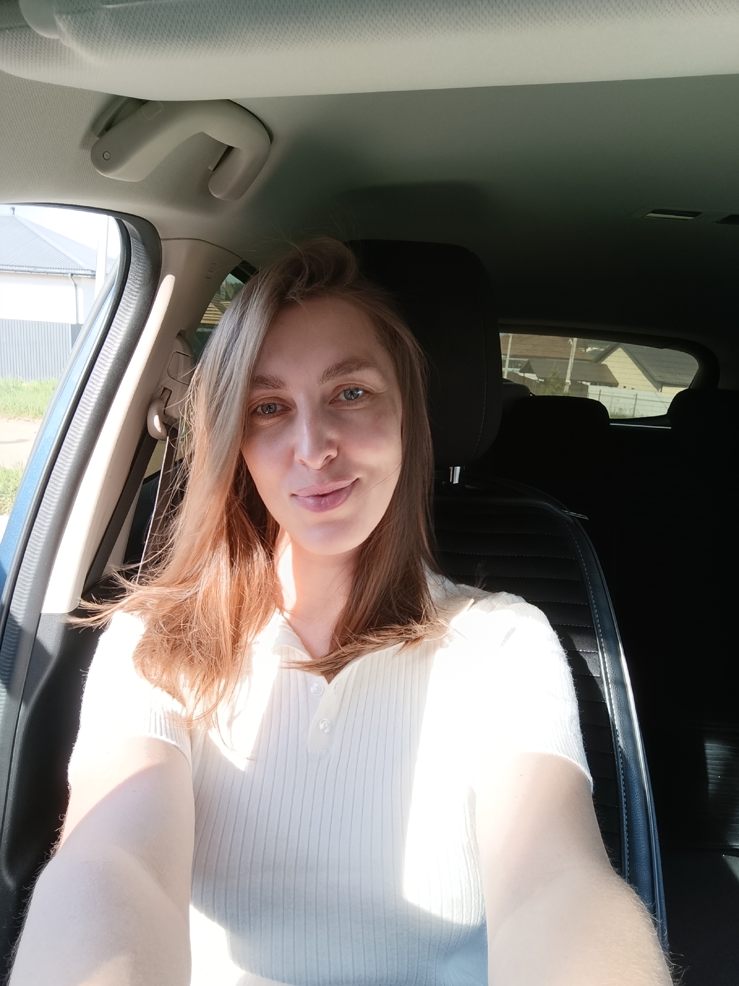 A woman with light skin and brown hair is seated in a car. She is wearing a white, ribbed shirt with buttons. The car's interior is visible, including the dashboard, passenger seat, and headrest. Outside the car window, blurred glimpses of houses and greenery suggest a residential area. Given the request for location details within the city of Chaikovsky, Russia, one can infer that the photo was taken in a residential area of that city. The exact street or address is impossible to determine without further information.