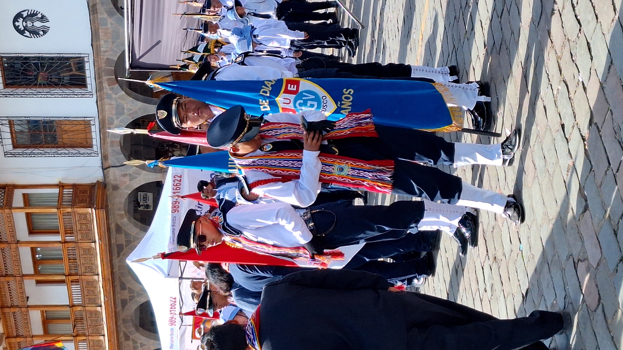 The image depicts a street scene in Cuzco, Peru, likely during a parade or official event. The foreground features several young men in uniform, standing on a cobblestone street. They are wearing dark blue pants, white shirts, black hats with gold trim, and sashes with colorful, traditional Andean patterns. Some are holding flags. In the background, a white tent is visible, with text reading "Peruano Suiza 989-316622." The architecture of the buildings behind the tent, with wooden balconies and arched windows, is characteristic of Cuzco's colonial style. The overall impression is one of a cultural or ceremonial gathering in a historic urban setting.
