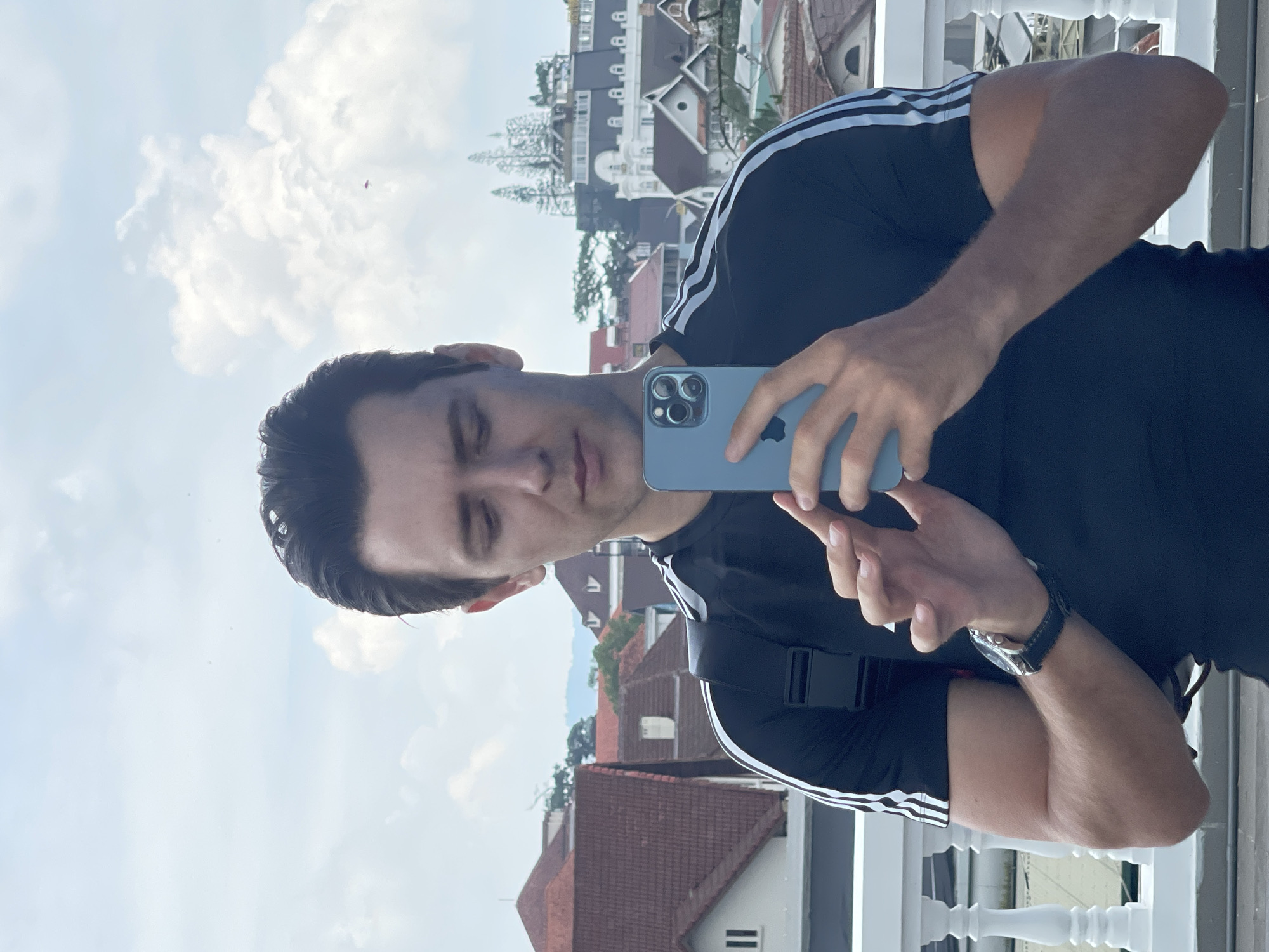 The image shows a man taking a selfie with a blue iPhone on a balcony. He is wearing a black t-shirt with white stripes on the sleeves and a black watch. In the background is a cityscape of Lâm Viên - Đà Lạt, Vietnam, with various buildings, including a large white one with multiple windows, visible under a cloudy sky.