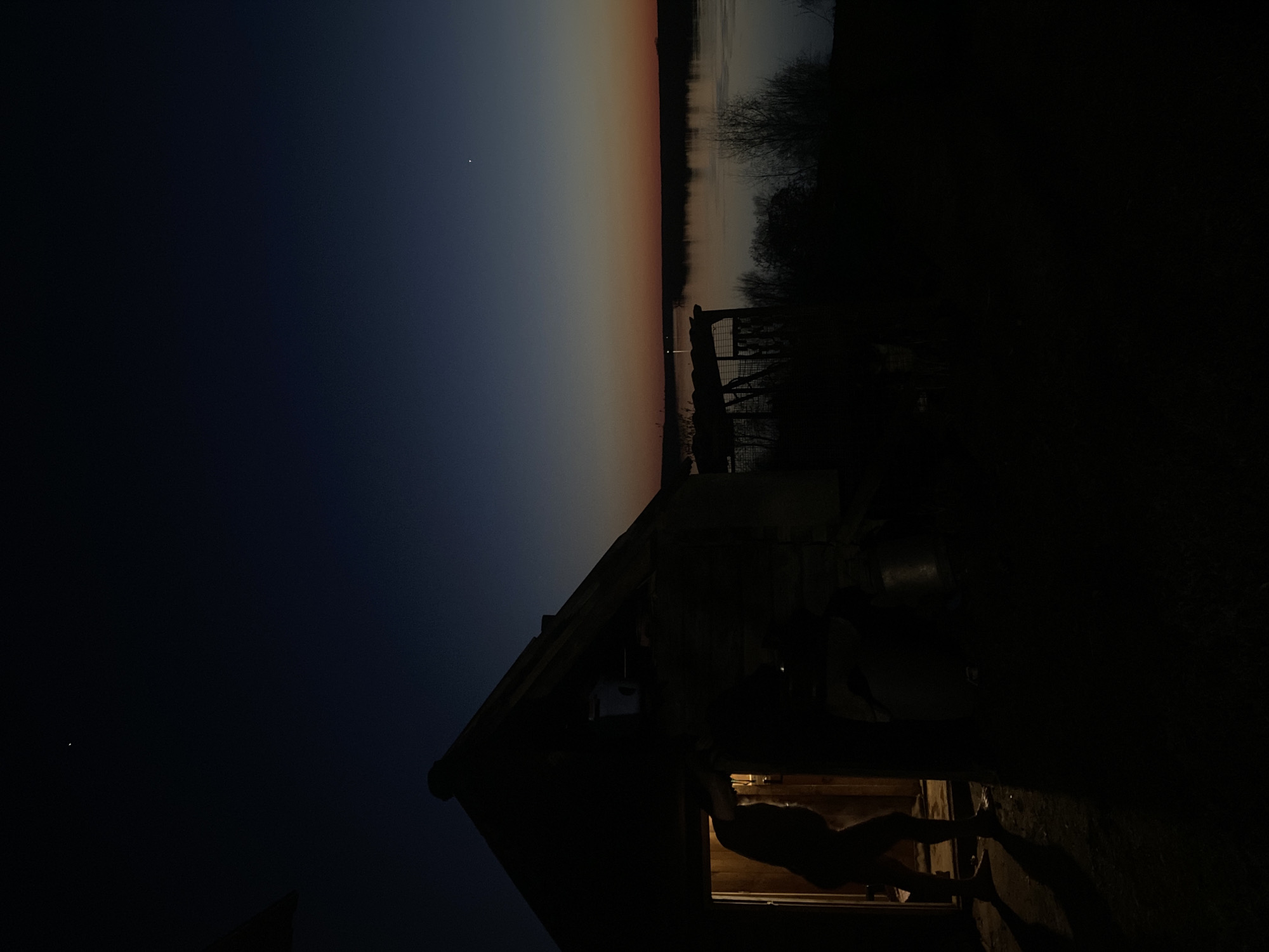 Captured at dusk in Ольховка, Russia, the image presents a silhouette of a building against a backdrop of a sky transitioning from dark blue to orange. The silhouette of a human figure is visible within the lit window of the structure. The background includes dark shapes indicating trees and possibly a body of water.