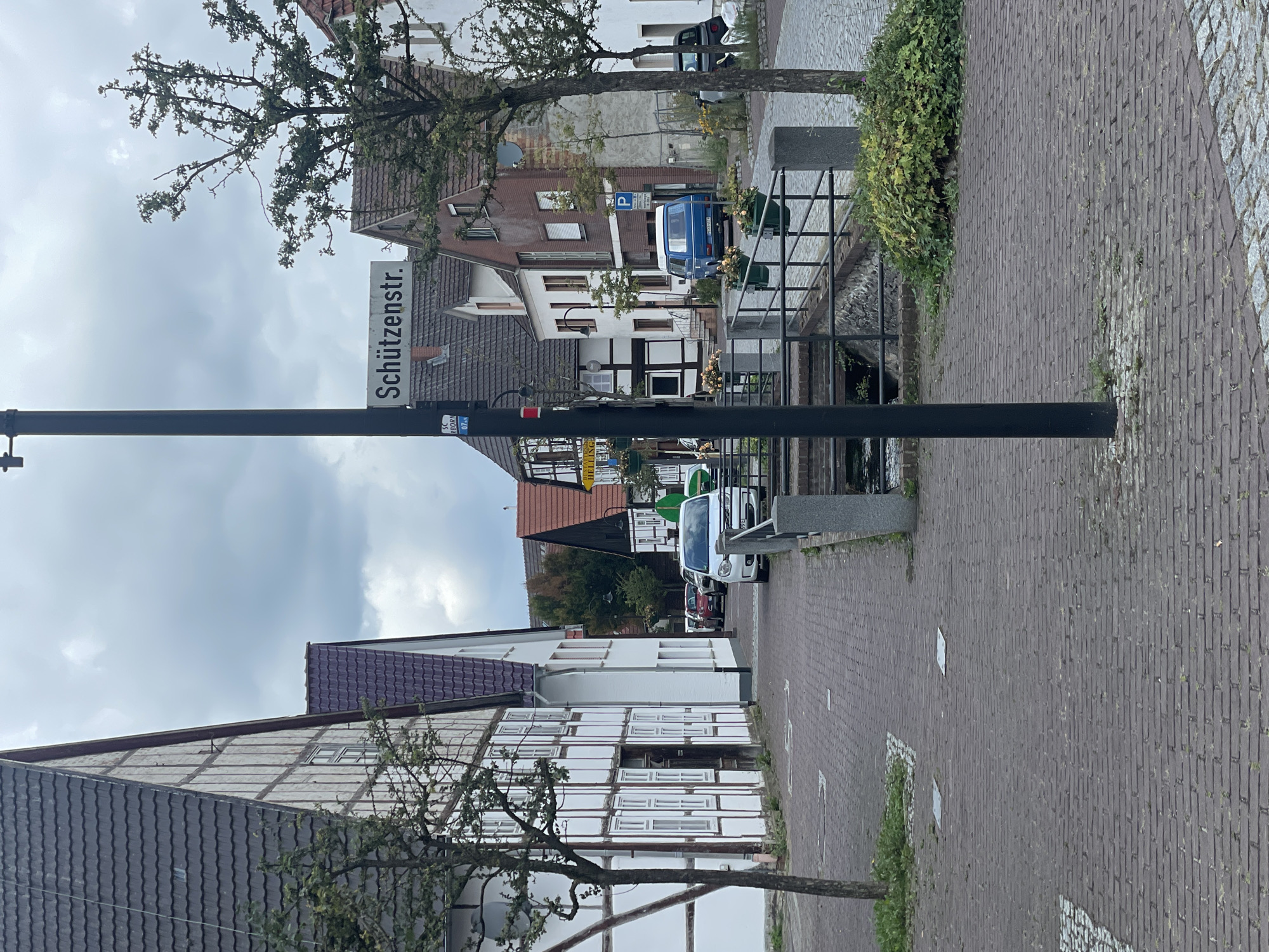 The image depicts a street scene in Salzkotten, Germany. A black lamppost with a street sign that reads "Schützenstr." stands prominently in the foreground, slightly off-center to the left. To the left of the lamppost is a traditional half-timbered building with a dark tiled roof and white infill between the timbers. Adjacent to this building, and further down the street, are other buildings with tiled roofs, including a red-brick building. A small canal runs alongside the paved walkway, bordered by a metal railing with planters. Several cars are parked on the street, including a blue van and a white minivan. The ground is paved with red bricks and cobbles. The sky is overcast with clouds. The overall impression is of a quiet, historic street in a German town.