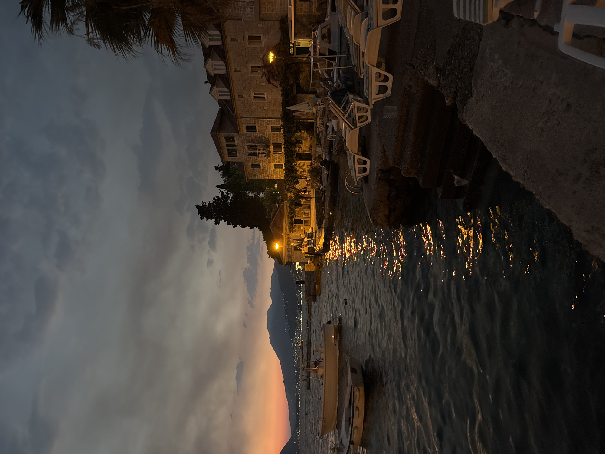 The image depicts a waterside scene at dusk. In the foreground, the dark water is gently rippling, reflecting the warm glow of lights from the shore and the fading sunset. Several white lounge chairs are arranged on a stone embankment to the right, some appearing empty.
Along the shore, a traditional stone building with multiple stories and illuminated windows is visible. A street lamp casts a bright yellow light near the building. Further back, more buildings are clustered, some with pitched roofs and dormer windows. Trees and foliage are interspersed throughout the scene.
Two small white boats are moored in the water to the left. In the background, a mountainous landscape is silhouetted against the colorful twilight sky, with distant lights suggesting a town or city. The sky itself is a mix of dark clouds and hues of orange and yellow from the setting sun. The overall atmosphere is serene and tranquil, capturing the transition from day to night.