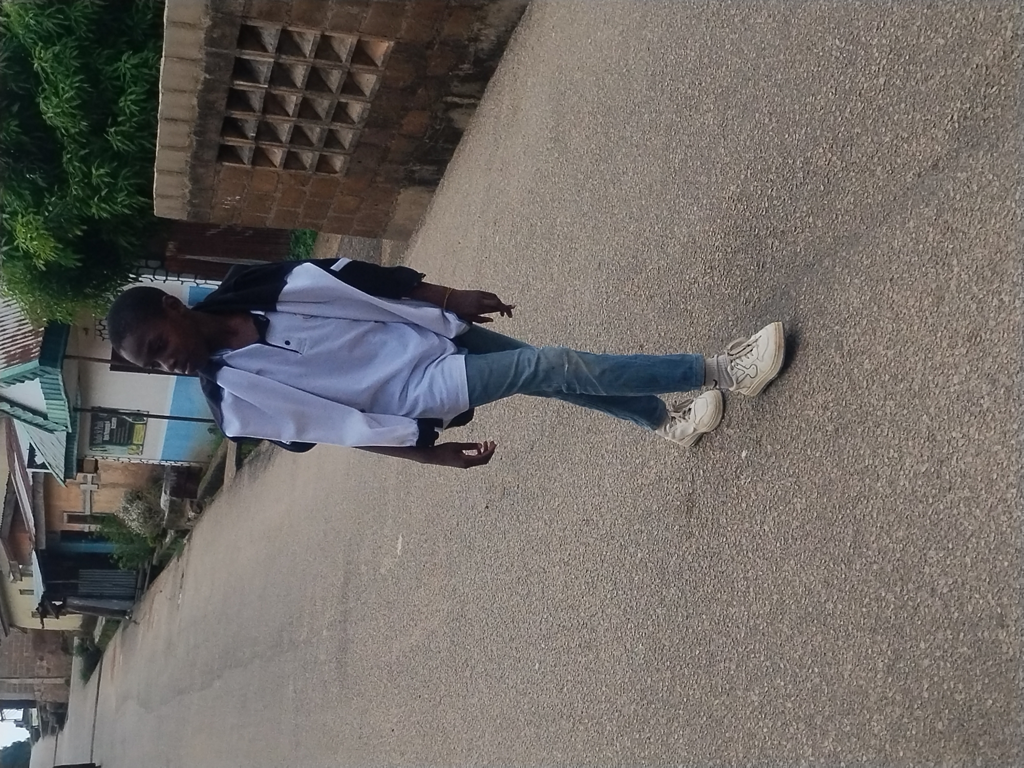 A young person, dressed in a white polo shirt, blue jeans, and white sneakers, stands on a paved road. They are wearing a black and white jacket draped over their shoulders. The person is looking down and to their left, with their right hand slightly extended.
The scene is set in Kabong, Nigeria. In the background, to the left of the person, is a building with a corrugated metal roof and some windows. Further back, there's a wall with a textured pattern and lush green foliage from a tree. The road itself is asphalt, with a light grey, speckled texture. The lighting suggests it is daytime, likely morning or afternoon, with no strong shadows indicating harsh midday sun. The overall atmosphere appears casual and everyday. No visible text is present in the image.
